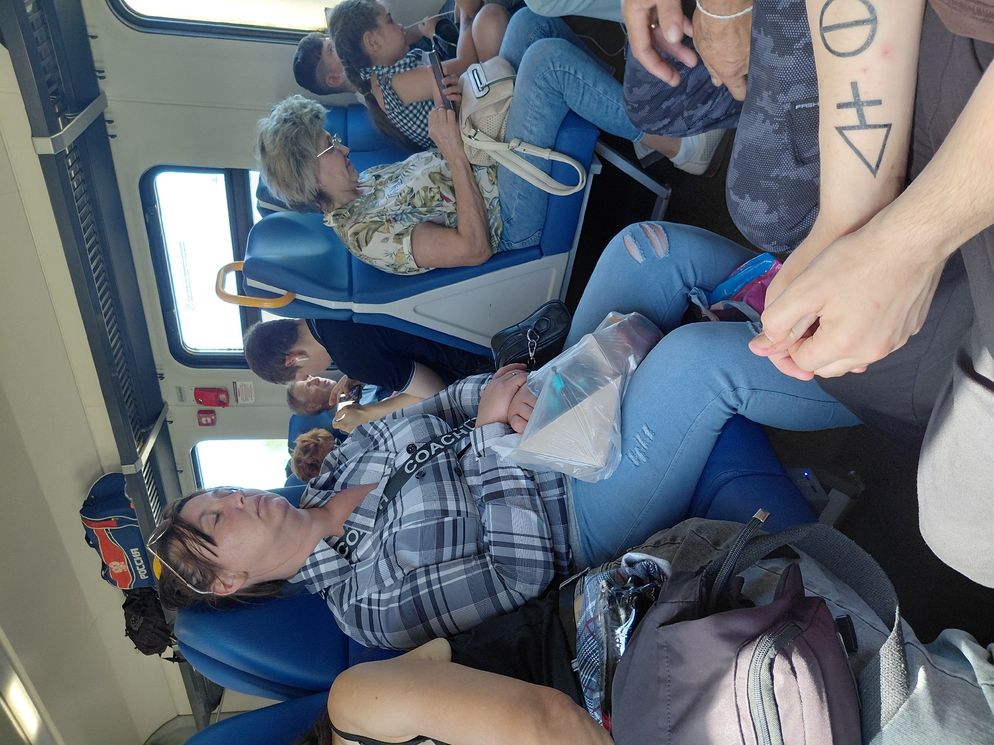 The image depicts the interior of a train carriage, possibly located in Novosibirsk, Russia, judging by the "РОССИЯ" (Russia) branding on a bag overhead. Several passengers are seated. In the foreground, a woman with her eyes closed appears to be sleeping, wearing a plaid shirt and blue jeans. She has a plastic bag containing an object on her lap. To her left, a portion of another passenger is visible, wearing a black tank top. To the right of the sleeping woman, an older woman is looking at her smartphone, wearing a floral patterned shirt and blue jeans, with a cream-colored purse on her lap. Further back, other passengers are partially visible. The train carriage is equipped with blue seats, overhead storage, and windows. The overall impression is of a typical train journey with passengers resting or engaging with their devices.