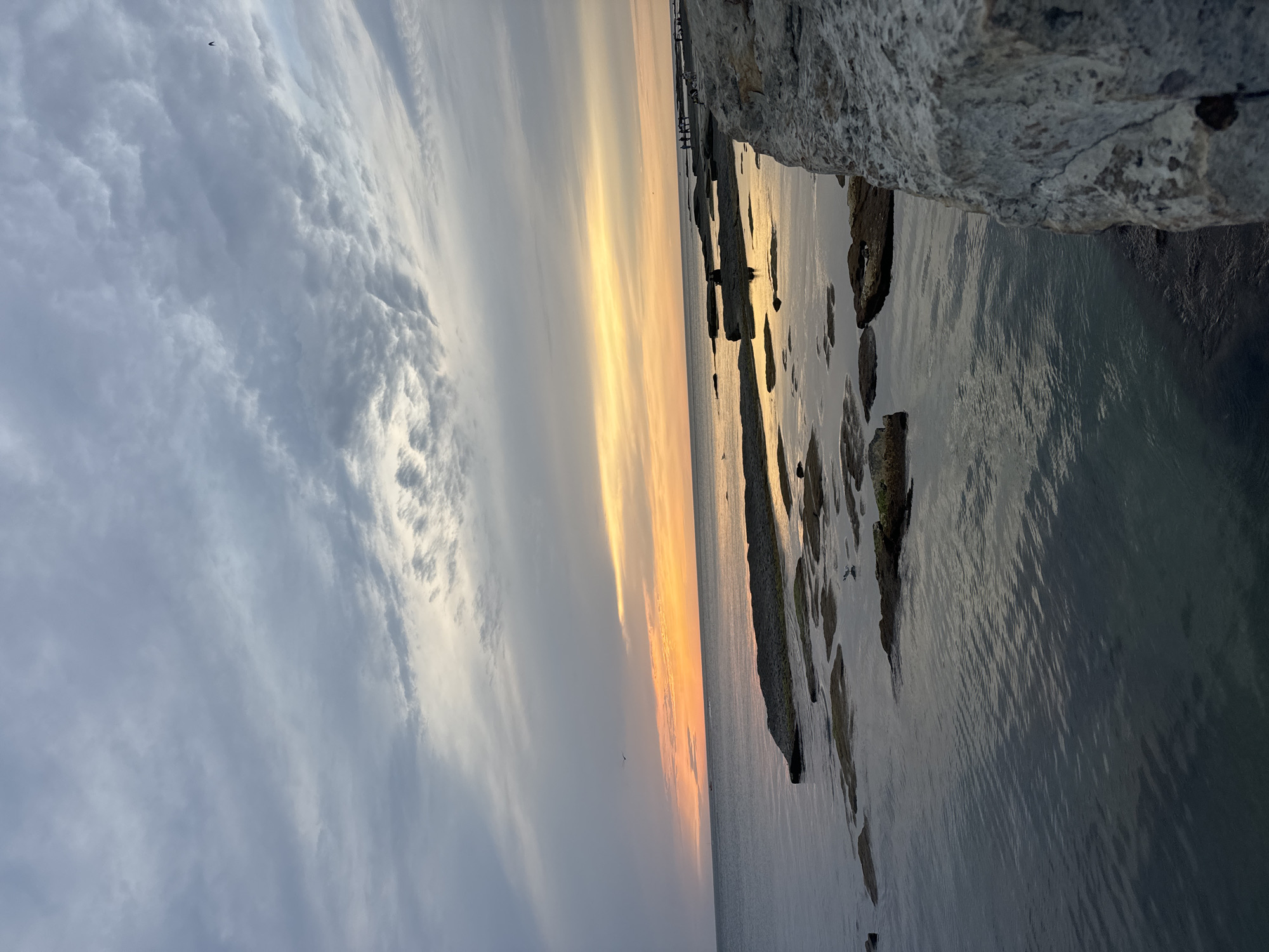 A seascape at sunset depicts the Caspian Sea under a cloudy sky. The water is calm, reflecting the muted orange and yellow hues of the setting sun near the horizon. Several rocky outcrops emerge from the water, some partially submerged. In the foreground, a large, textured rock formation occupies the right side of the frame. Along the distant shoreline, a line of people are visible, appearing as small silhouettes. A few birds are in flight in the sky. The scene captures a tranquil moment at sunset along the coast of Aktau, Kazakhstan.