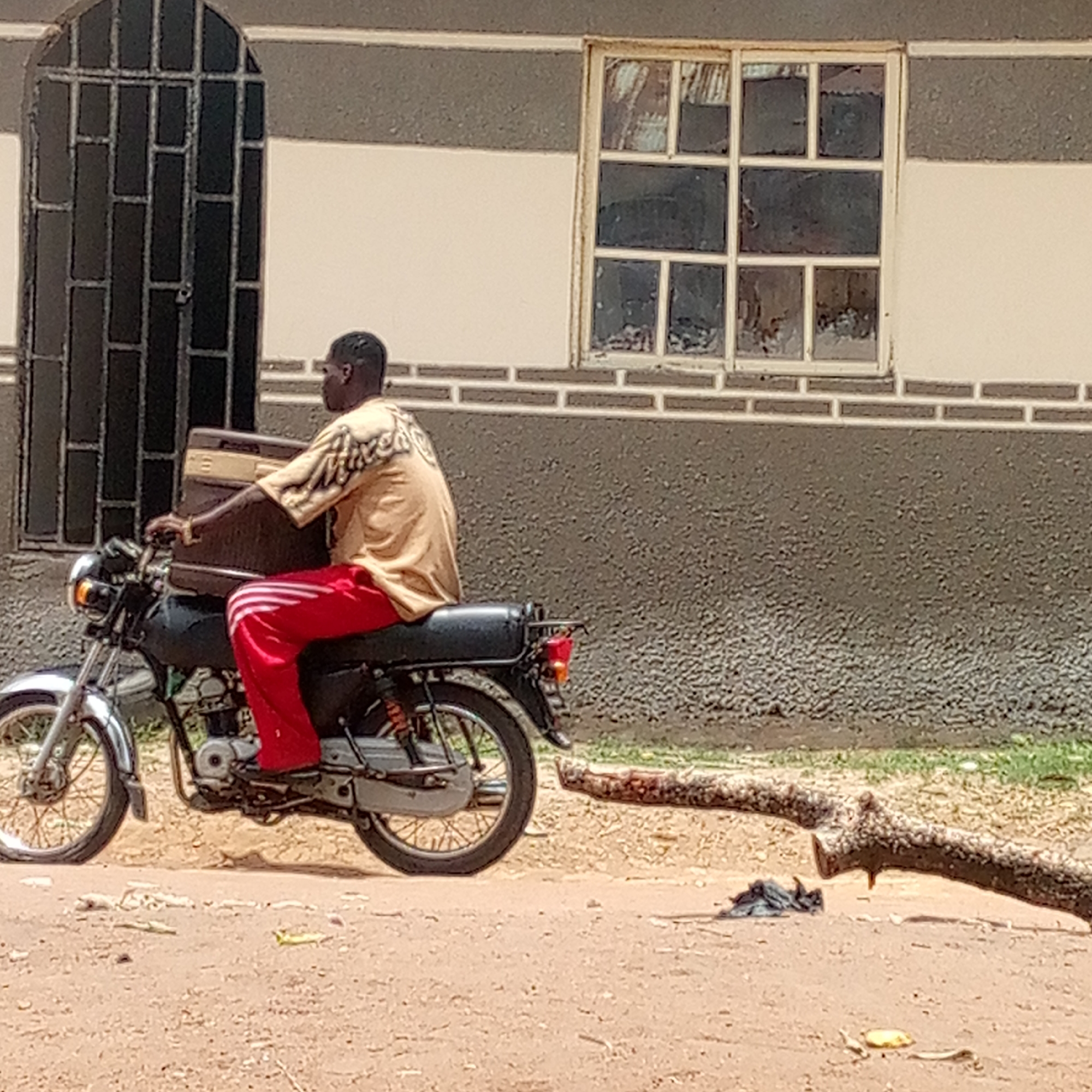 A man rides a black motorcycle on a dirt road in an unspecified city in Unknown. He wears a tan patterned shirt and red sweatpants with white stripes. He carries a brown rectangular object on the motorcycle's gas tank. A fallen tree branch lies on the right side of the road. Behind the man is a two-story building with a dark grey base, a light tan upper portion, and a dark grey door and window with geometric patterns.
