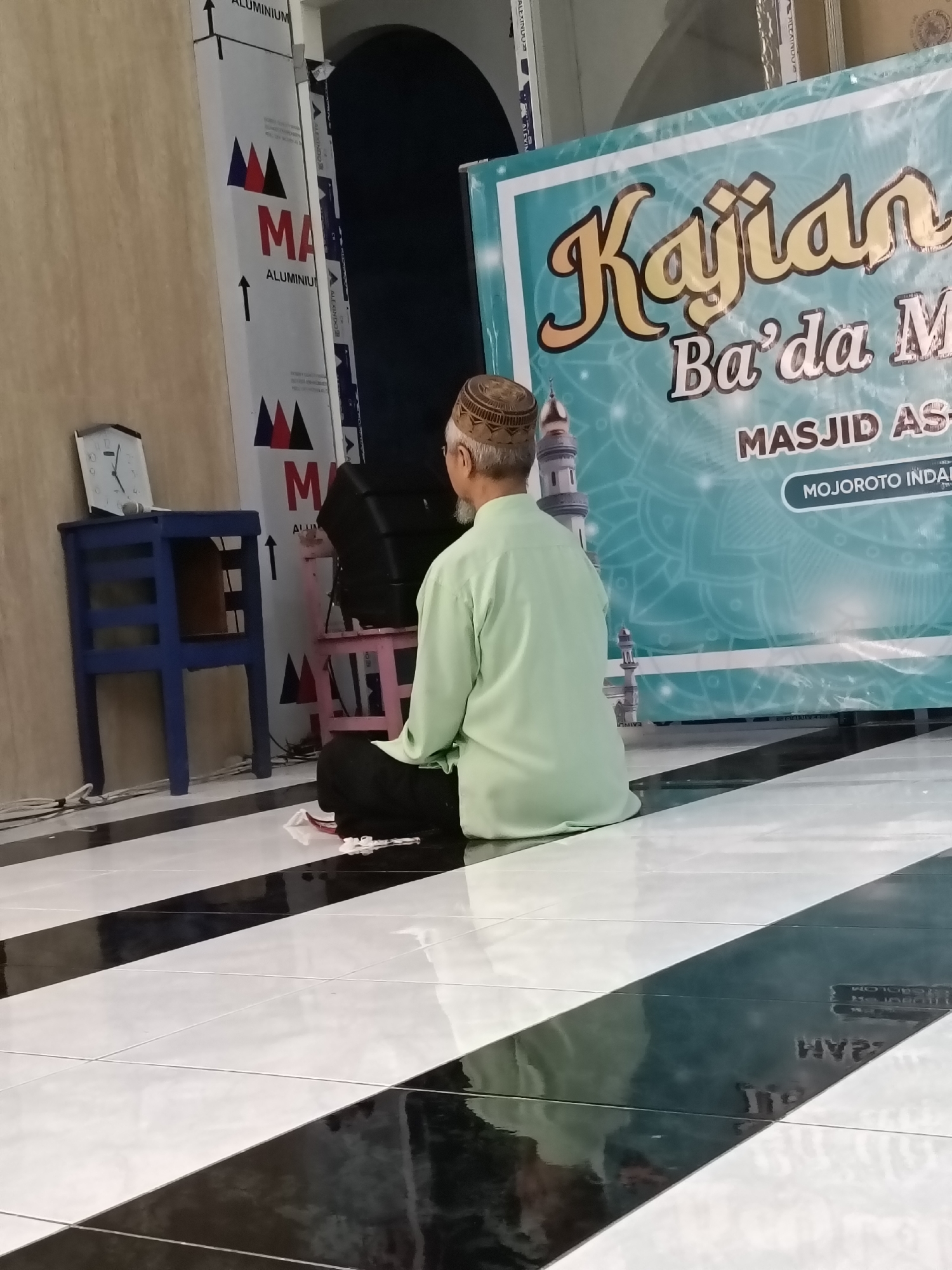 In a room with a tiled floor featuring a bold black and white diagonal pattern, a man sits facing away from the camera, dressed in a light green tunic and a patterned kufi. He appears to be seated on the floor in front of a large banner. The banner, predominantly teal and white with intricate mandala-like designs, displays text in Indonesian. Prominently featured are the words "Kajian Ba'da M" in a stylized gold font, followed by "MASJID AS-" and then "MOJOROTO INDAH" on a white curved banner. To the left of the man, a small blue wooden table holds a clock showing approximately 9:10. Behind the table and to the left of the man is a tall, metallic structure with "MA ALUMINIUM" printed on it. A black speaker sits on a small pink stool to the right of the man. The overall scene suggests an indoor event or gathering at the Majestik Asal Mojoroto Indah mosque in Kediri City, Indonesia.