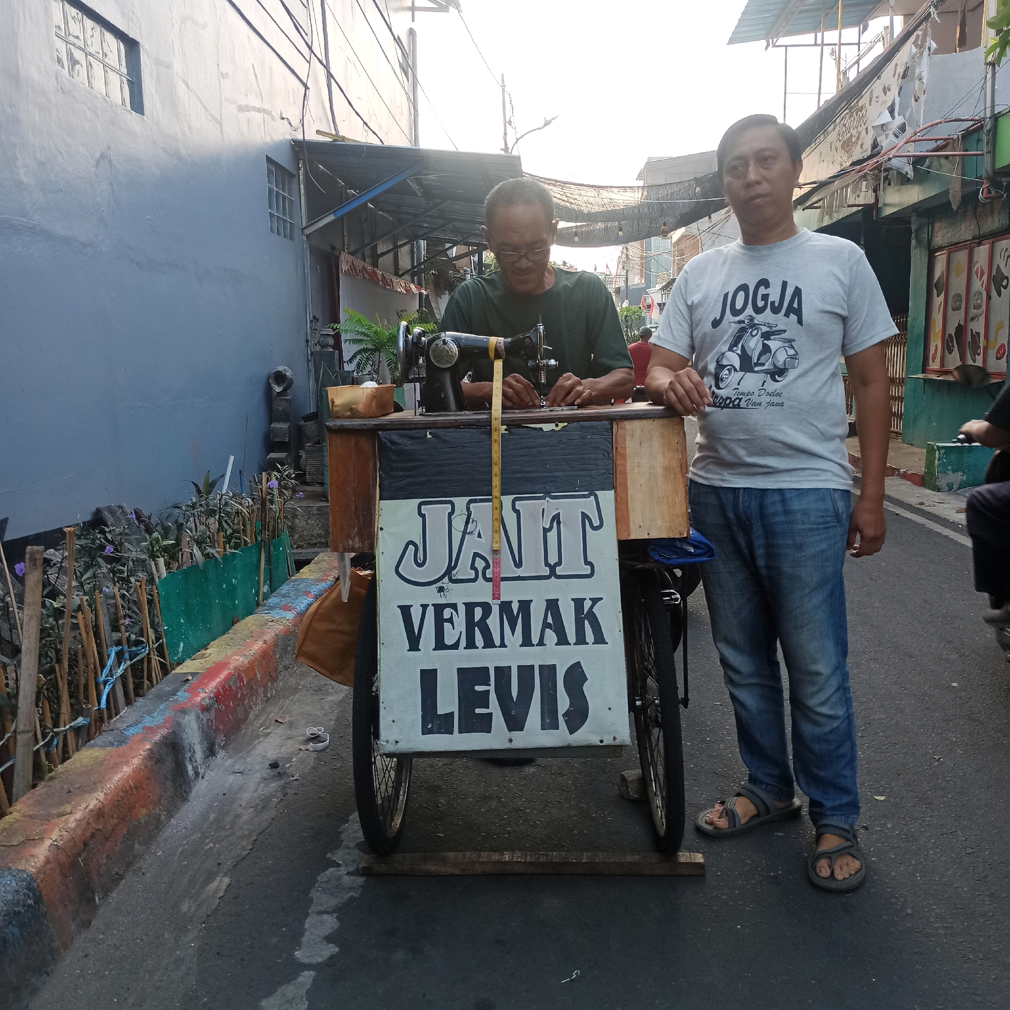 Two adult males are standing on a paved street next to a bicycle-powered sewing cart. The cart has a sign on the front that reads "Jahit Vermak Levis," which translates to "Sewing and Alteration for Levi's." The man on the left, wearing a dark green t-shirt and glasses, is seated and operating a vintage sewing machine. The man on the right, wearing a light grey t-shirt with a graphic of a scooter and the word "JOGJA," is standing and looking towards the camera. The environment appears to be an urban setting with buildings in the background and a sidewalk with some greenery along the left side of the street. The lighting suggests it is late afternoon or early morning. The image captures a scene of a mobile tailor at work in a residential or commercial area.