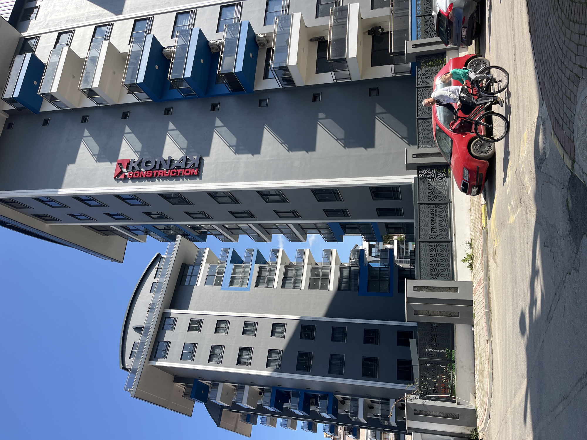 A medium shot captures a tall, gray-blue and white building in Alanya, Turkey, with the brand name "KONAK CONSTRUCTION" displayed in red lettering on the facade. A red car is parked in front of the building. A man in a white shirt and black pants stands next to the car, with a bicycle resting on the roof. A second, smaller person in a green shirt is partially visible in the driver's seat. The scene is set outdoors on a sunny day, with blue sky visible. A section of the street is also in view.