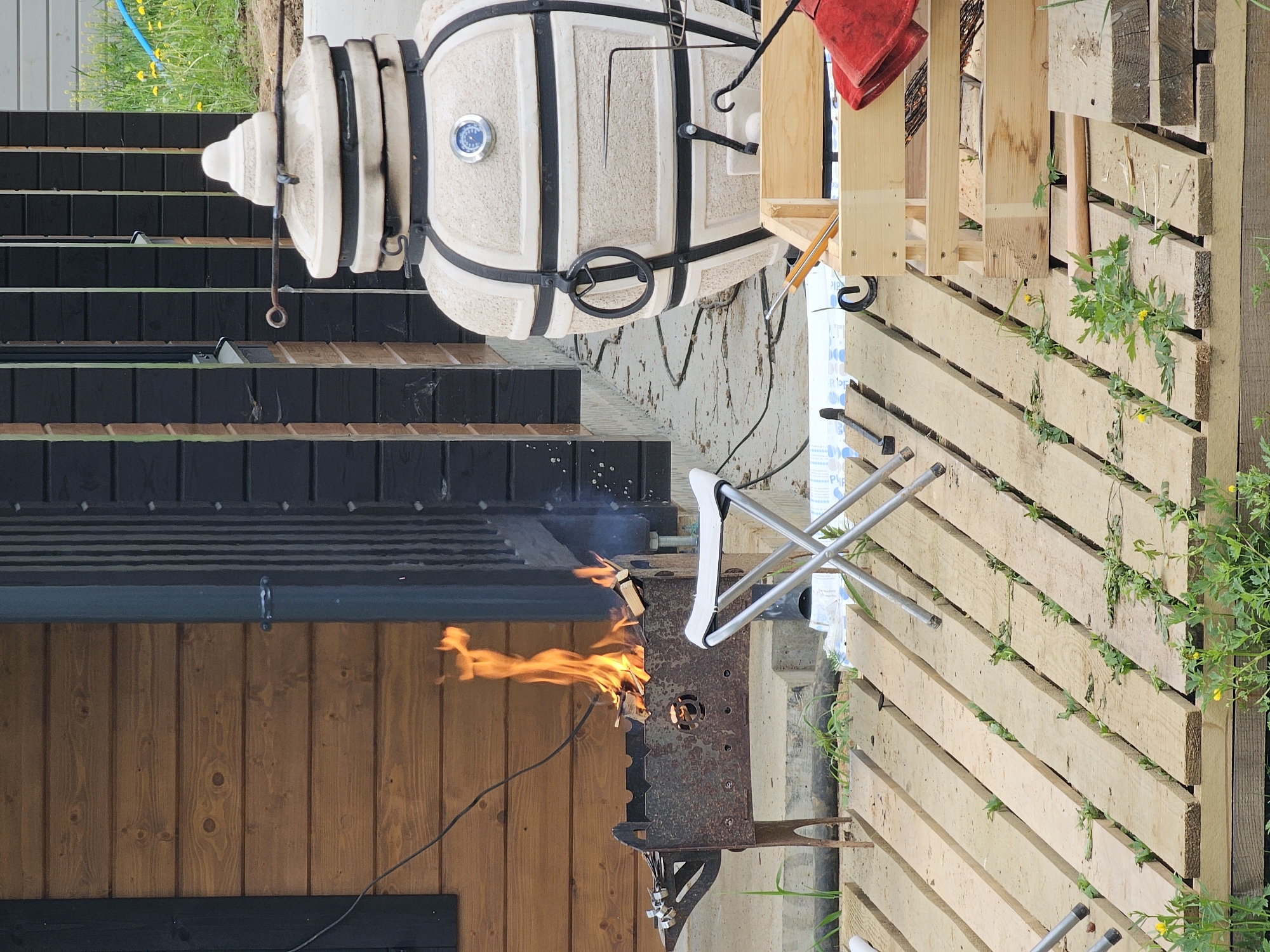 The image shows an outdoor setting, likely in Мышкино, Russia, given the prompt context. The scene includes a wooden building with vertical planks, a wood-burning stove with flames visible, and a ceramic tandoor oven. A wooden pallet stands adjacent to the stove. The scene also features metal and plastic objects, including a folding chair, and some vegetation growing between the wooden planks of the pallet. No people are visible in the image.