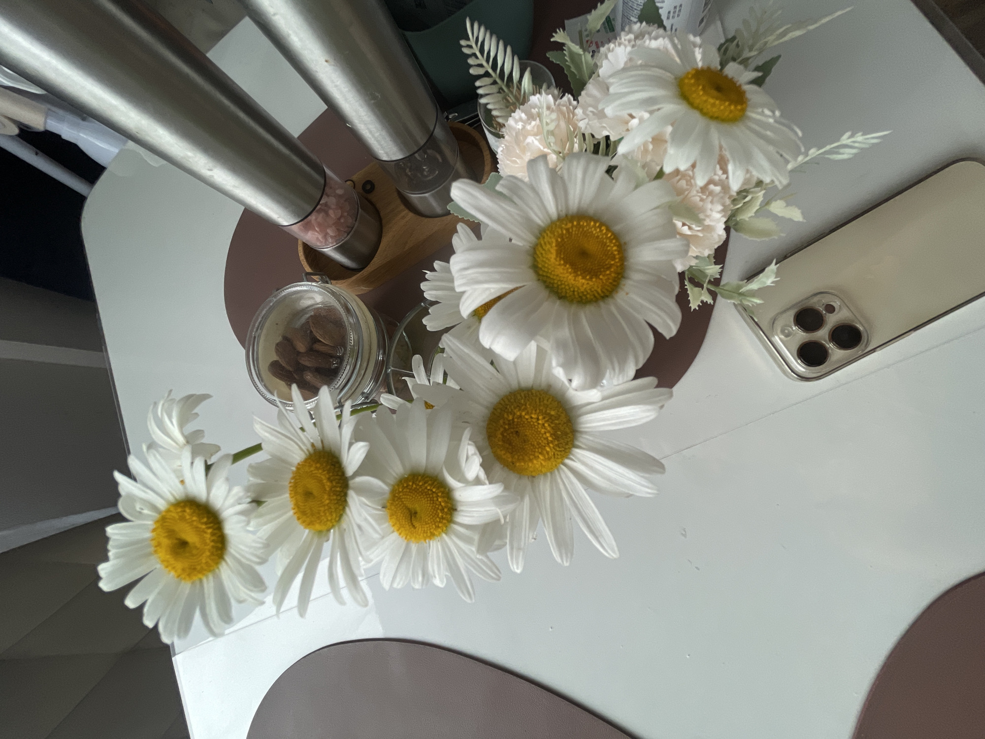 A close-up, top-down shot features a cluster of white daisies with yellow centers arranged on a white table. To the right, a wooden holder contains two stainless steel grinders, one filled with pink salt crystals, and a small glass jar of almonds. In the foreground, a light-colored smartphone with a triple camera system rests on the table. The background is blurred, suggesting an indoor setting. The composition is angled, with a portion of a dark brown placemat visible on the left. The image captures a still-life arrangement, devoid of any human presence or discernible urban context that would place it in Подшивалово, Russia.