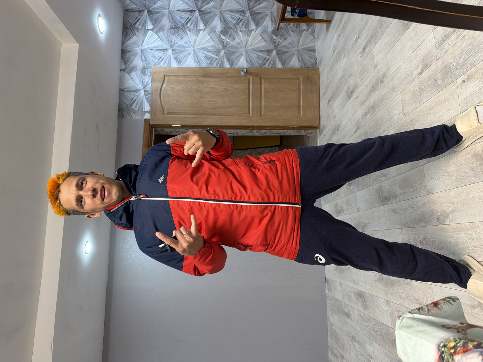 An energetic man poses indoors, rotated 90 degrees clockwise in the image. He has distinctive bright orange, spiky hair and a wide smile, displaying a playful expression. He is wearing a two-tone hooded jacket, predominantly red on the torso with navy blue sleeves and hood, featuring a white stripe across the chest and an "M" shaped logo (likely Quechua) on the left chest area. His lower attire consists of navy blue sweatpants with a white "e" logo (consistent with ASICS) on the left thigh and light-colored sneakers. His left hand is raised, making a "rock on" gesture with an apparent ring on his ring finger. His right hand also makes a gesture, with the index finger pointing outwards and the thumb extended (shaka sign), and a watch is visible on his right wrist.
The setting appears to be a room or hallway in Chaikovsky, Russia. The background features a plain light-colored wall on the left, and a more decorative wall with a modern, geometric, fan-like pattern in shades of gray and white. A light brown wooden door with paneling is visible in the upper background. The floor is covered with light-colored laminate or wood-look flooring laid in planks. Two bright, circular recessed lights illuminate the ceiling. Partially visible in the bottom right corner is a patterned object, possibly a gift bag or box, decorated with a deer motif and festive elements.
