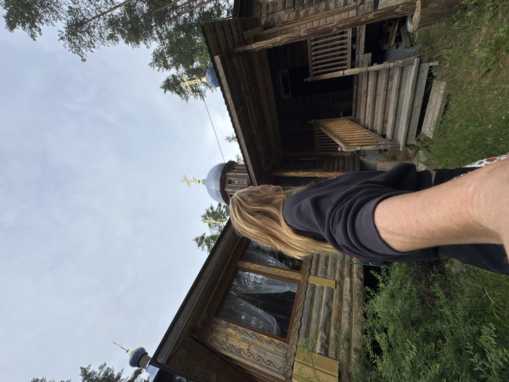 The image shows a person taking a selfie in front of a wooden Orthodox church in Nenimyakii, Russia. The church has a domed roof with a cross on top, and it is surrounded by trees. The person is wearing a black sweatshirt and has long blonde hair. The sky is overcast. The church appears to be made of logs, with intricate carvings around the windows. There is a wooden porch with stairs leading up to the entrance. The area around the church is covered in grass and greenery.