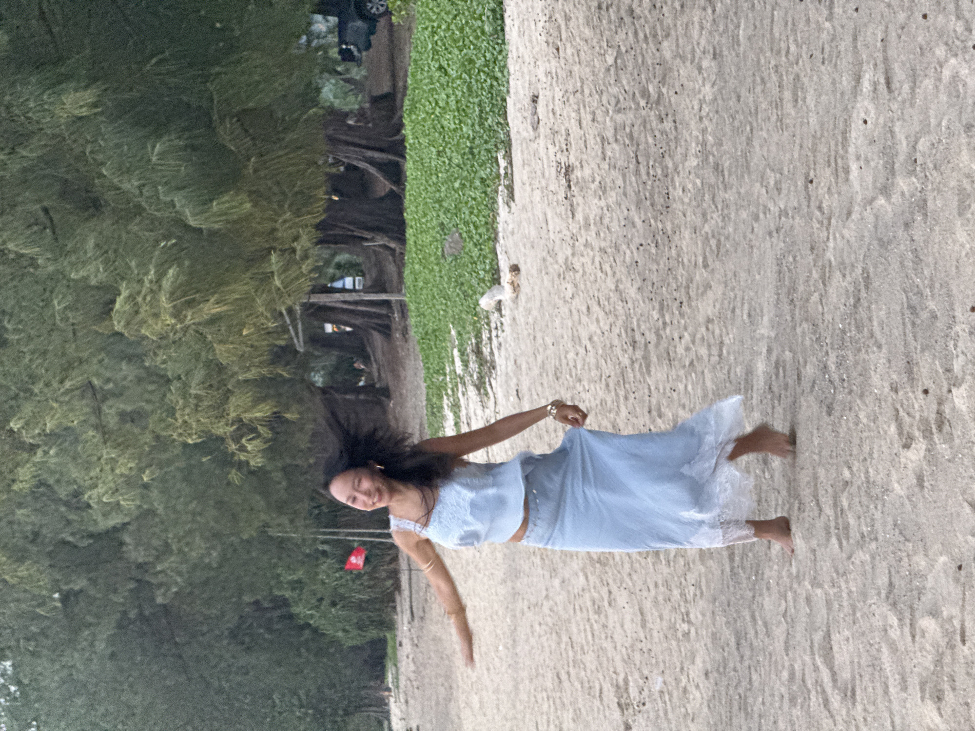 A woman is walking on a sandy beach. She is wearing a light blue, two-piece outfit with lace trim on the skirt. Her arms are extended, and her hair is blowing in the wind. Behind her, there are trees and vegetation. The lighting suggests it might be late afternoon or early evening.