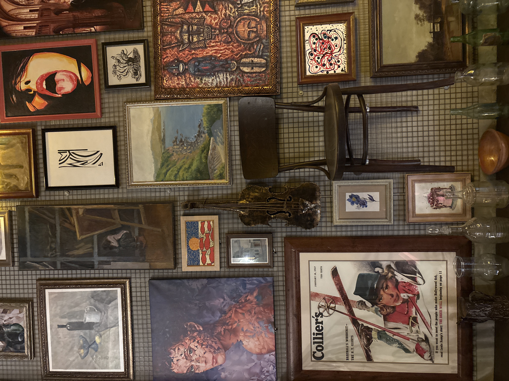 The image depicts a wall in Kazan, Russia, densely decorated with a variety of framed artworks, including paintings, drawings, and a vintage magazine cover ("Collier's," January 18, 1947). An upside-down wooden chair is mounted on the wall alongside the artworks. A sculpted golden violin, several antique glass bottles, and other decorative objects are integrated into the composition. The wall itself appears to be a wire grid, adding a structural backdrop to the eclectic display.