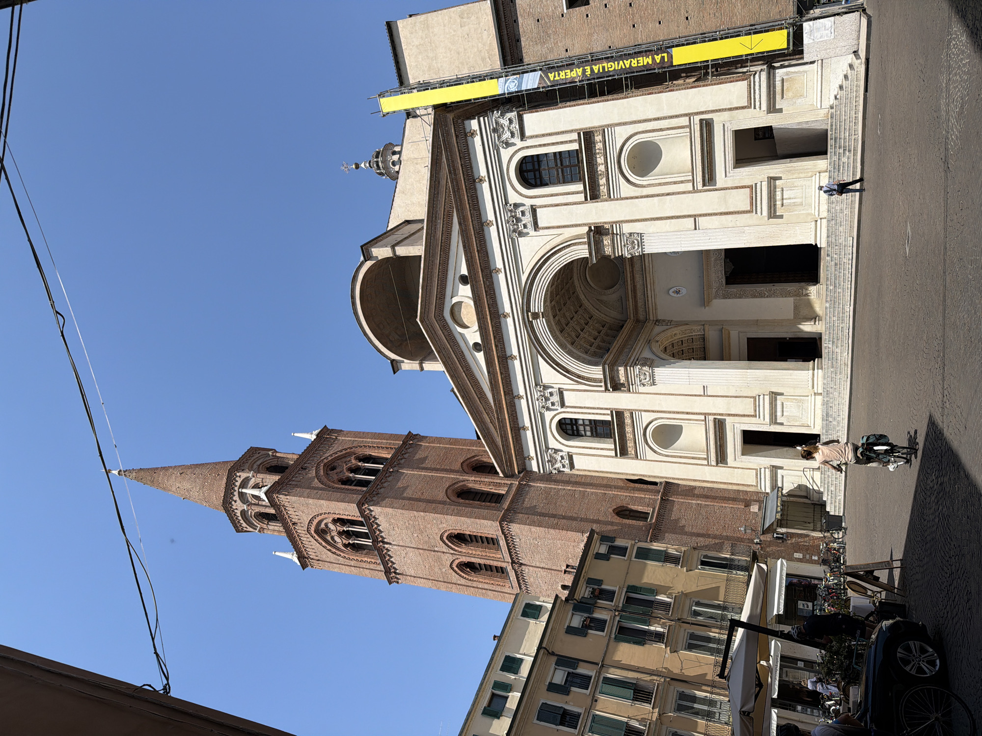 The image captures a view of Mantua, Italy, specifically highlighting the Basilica di Sant'Andrea. The facade of the basilica is predominantly cream-colored with intricate architectural details, including archways, columns, and windows. A yellow banner with Italian text ("LA MERAVIGLIA È APERTA") is positioned above the facade. To the left of the basilica, a brick bell tower with pointed spires rises prominently. Several utility wires cut across the upper left portion of the frame.
At street level, a person is walking by with a bicycle in front of the basilica. The street scene includes parked bicycles, an awning, and the front of a parked car. The light suggests it is daytime. The sky is a clear blue, and the architecture is lit by the sun.
The photo is taken from a low angle, emphasizing the height of the Basilica and the bell tower.