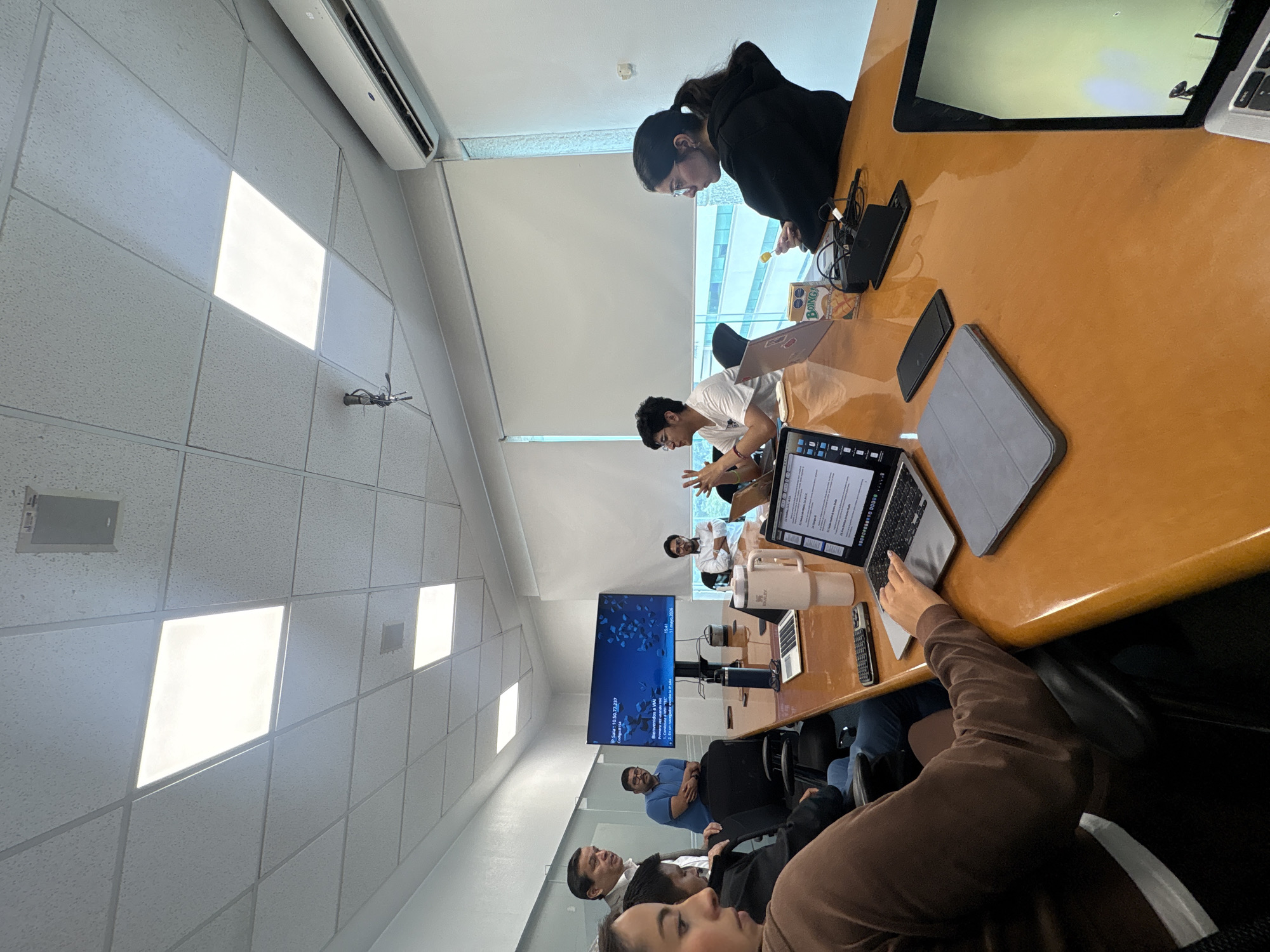 The image captures a meeting in progress around a large, orange-colored conference table in what appears to be an office setting. Multiple individuals are present; some are actively engaged with laptops and tablets, while others are seated and appear to be participating in the discussion. The presence of a display screen in the background suggests a presentation or video conference is taking place. The room has a drop ceiling with recessed lighting and windows providing natural light. The location is in the city of Puebla, Mexico, inferred from the information displayed on the screen.