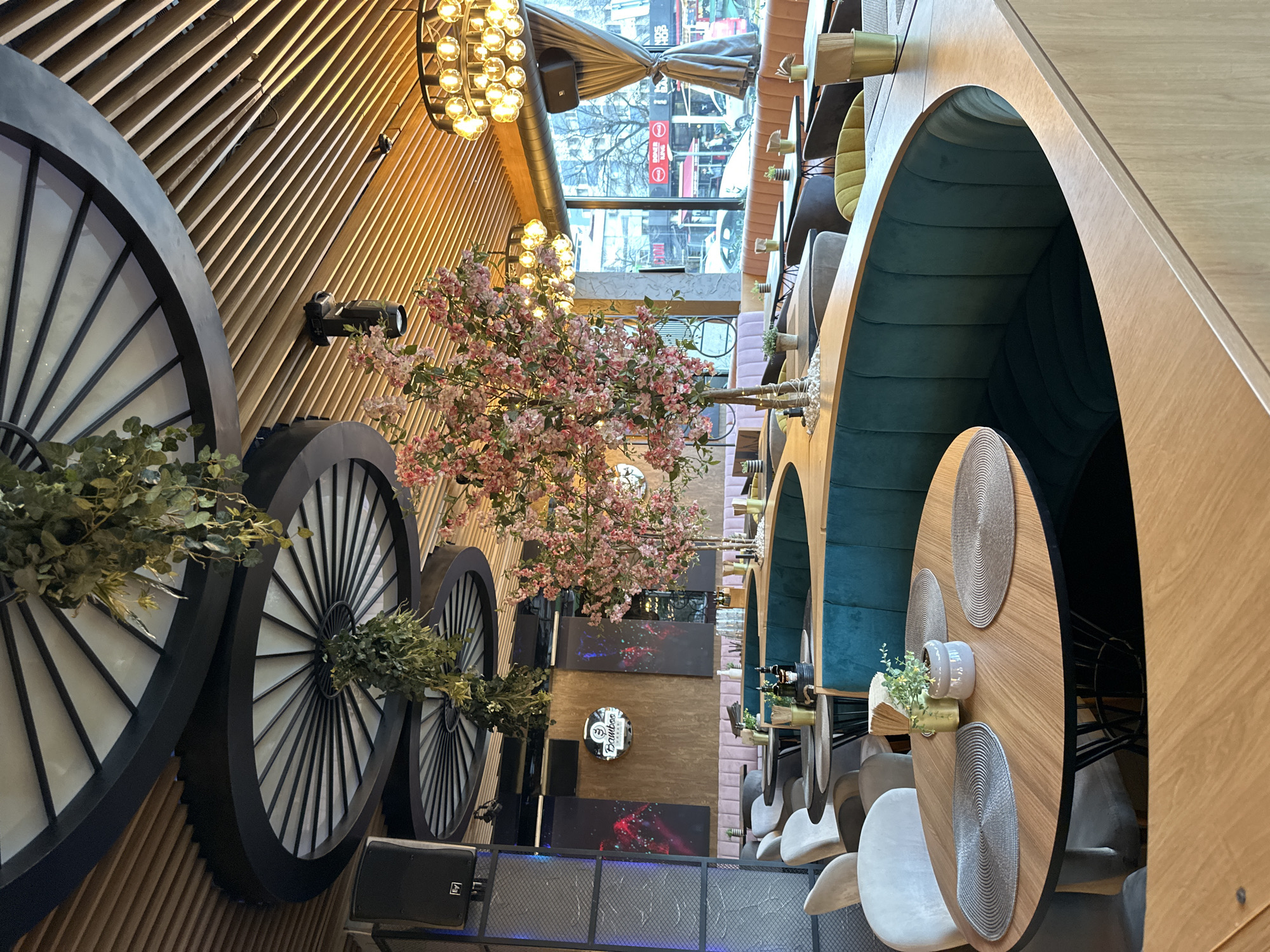The image depicts the interior of a restaurant in Sofia, Bulgaria. The perspective is from a high vantage point, looking down into the space. The restaurant features a modern design with elements such as large, decorative wheels affixed to a wood-paneled wall. Artificial flowering trees are placed along the wall. Dining areas include arched booths upholstered in dark green fabric, with round tables set with placemats and small decorative centerpieces. Chairs are upholstered in a light color. A window in the background offers a view of the street outside. The decor includes modern light fixtures. A metal cage is shown at the bottom of the image, which might be a stage or decorative feature.