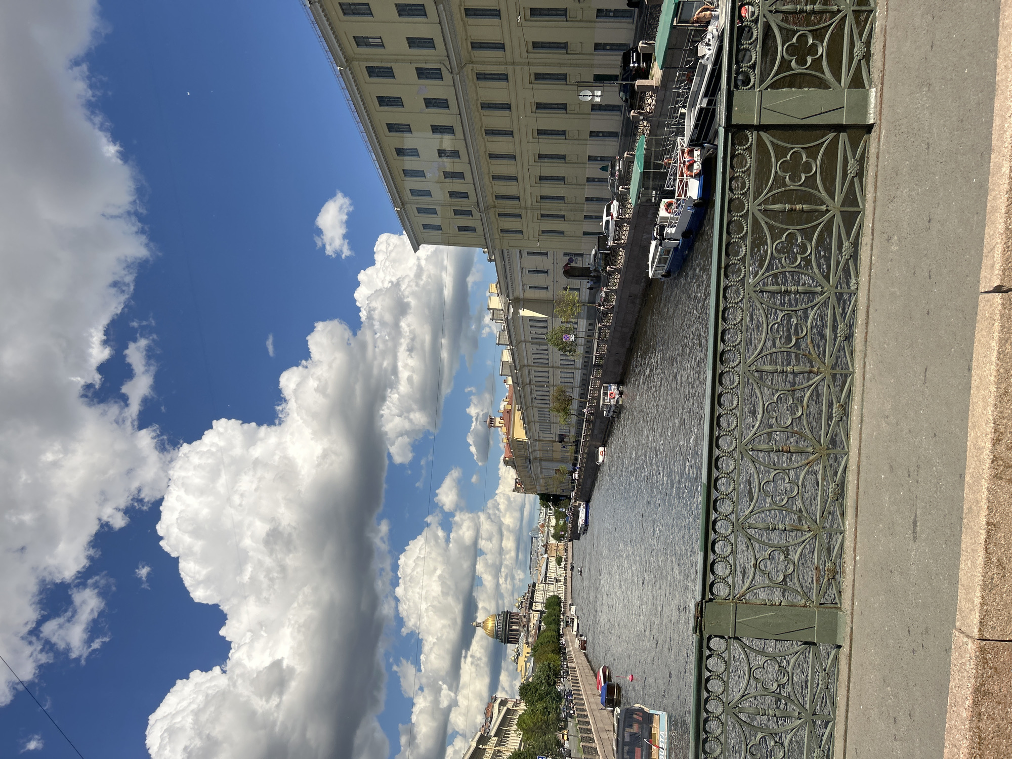 A long shot captures a view of a wide canal in St. Petersburg, Russia, under a bright, partially cloudy sky. The canal is lined with ornate, light-colored buildings on both sides. Numerous boats are docked or cruising on the water, and a large, golden-domed building, likely St. Isaac's Cathedral, is visible in the distance on the left bank. A detailed green metal railing runs along the foreground, separating the viewer from the canal. The water reflects the sky and surrounding architecture. The scene appears peaceful, with no discernable activity beyond the presence of the boats and the buildings.
