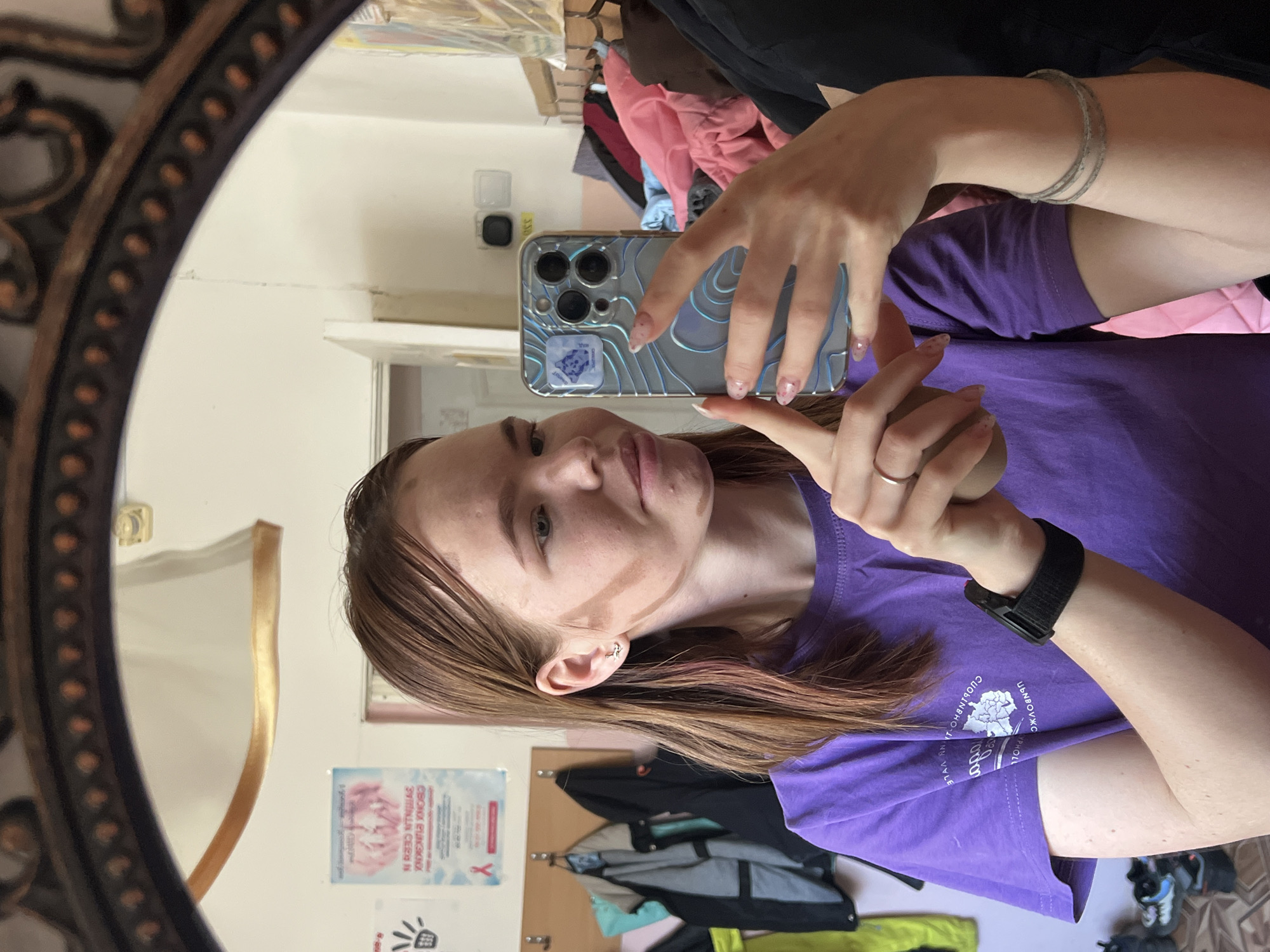 A person wearing a purple t-shirt takes a selfie in a mirror. The person is holding a phone with a patterned case. A silver bracelet is on the person's left wrist and a black watch band is on the right wrist. The mirror has an ornate gold and brown frame. In the background, there is a wardrobe, a clothes rack with clothes hanging on it, and various posters on the wall. The setting appears to be an indoor room. It is unknown which city the picture was taken.