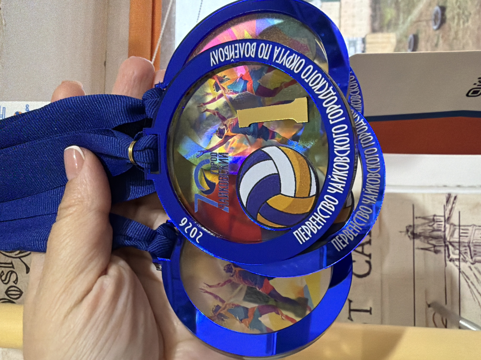 A close-up shot shows a fair-skinned hand holding two blue medals. The medals feature the text "ПЕРВЕНСТВО ЧАЙКОВСКОГО ГОРОДСКОГО ОКРУГА ПО ВОЛЕЙБОЛУ", which translates to "Championship of the Chaikovsky City District in Volleyball," indicating they are related to volleyball competitions in the city of Chaikovsky, Russia. The medals show images of volleyball players and a volleyball.