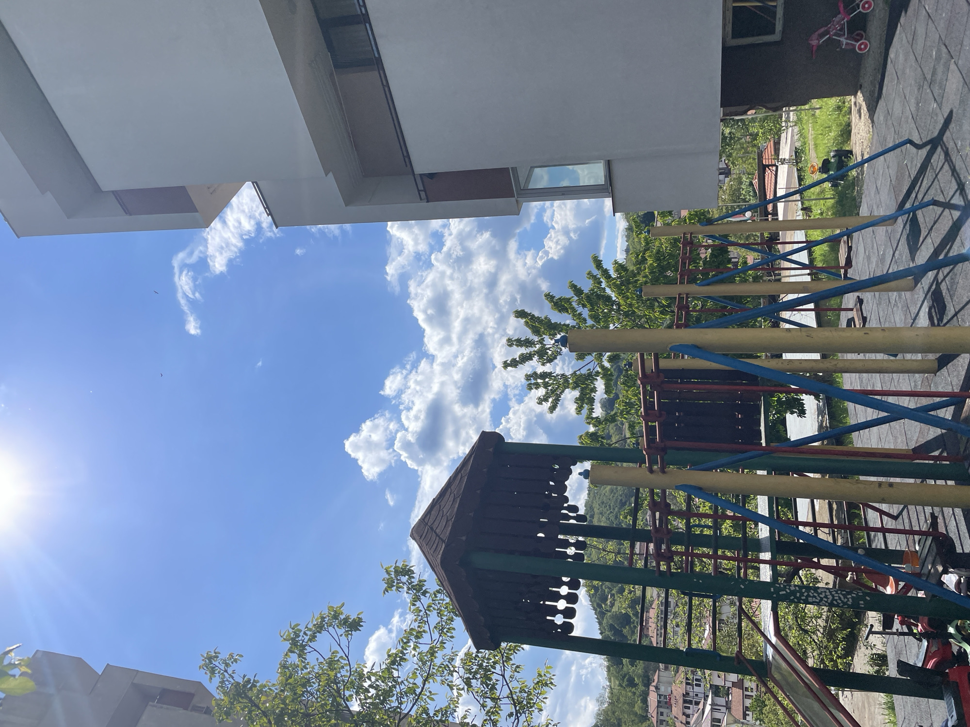 The image depicts a playground in Zlatograd, Bulgaria, viewed from a low angle, with a mix of architectural and natural elements. A portion of a white multi-story building is visible at the top left, contrasted by a bright blue sky dotted with cumulus clouds. The playground equipment includes metal climbing structures with yellow, blue, green, and red bars. A dark brown wooden playhouse roof is positioned in the lower center of the frame, partially obscured by tree branches with green leaves. In the background, a cityscape of Zlatograd is visible, with buildings interspersed with trees on a hillside. The image suggests a sunny day in a residential area of Zlatograd.