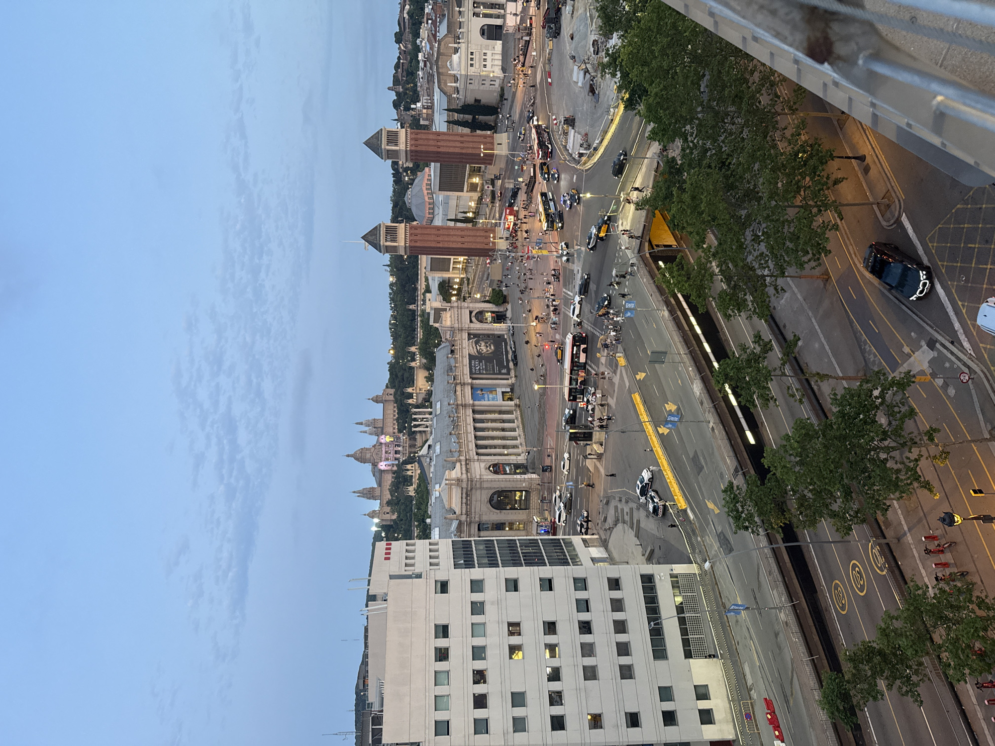 This is an aerial view of the Plaça d'Espanya in Barcelona, Spain, taken at dusk. In the foreground, a modern, multi-story white building is visible on the left. Below it, a two-level roadway system is active with vehicles, including cars and buses. Yellow lane markings and speed limit symbols are visible on the road surface. Trees line the lower roadway. In the middle ground, the iconic twin towers of the Venetian Towers stand prominently, flanking the Palau Nacional in the background. The Palau Nacional, a grand palace, is illuminated with lights. The sky is a pale blue with scattered clouds. The area appears to be a busy urban environment with many pedestrians visible on the sidewalks and crossing streets.