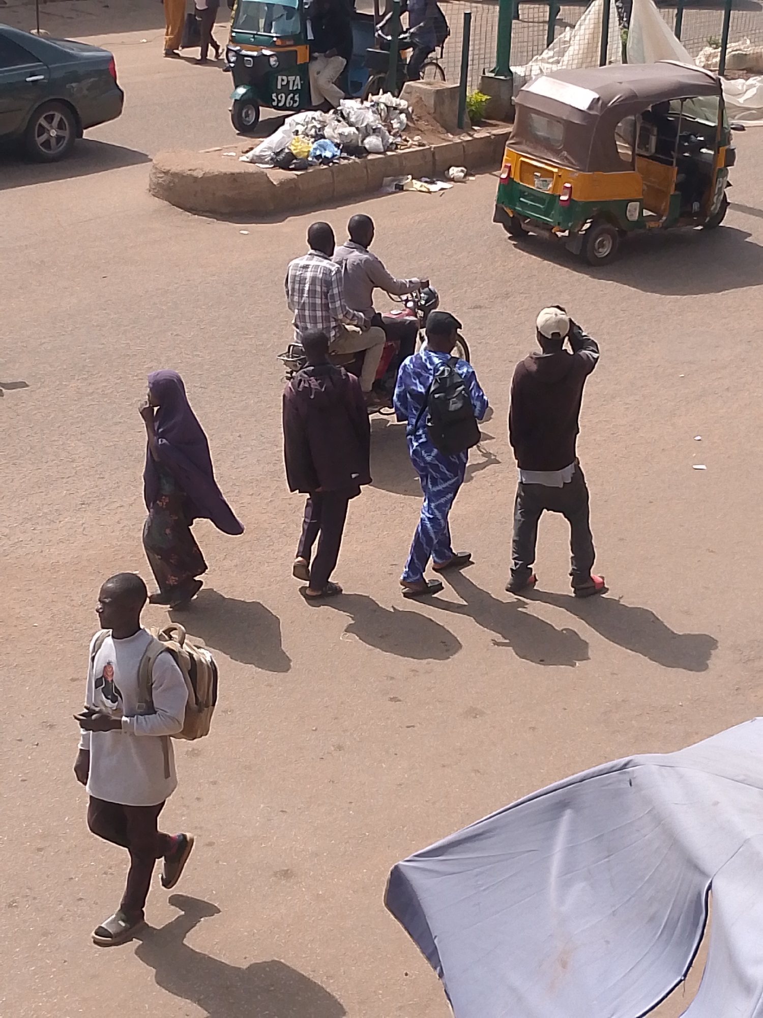 This image depicts a street scene in Bukuru, Nigeria. Several individuals are visible, walking and interacting within a dusty, outdoor environment, suggesting a warm and sunny time of day.
In the foreground, a young man wearing a white long-sleeved shirt with a graphic print and dark trousers walks to the left, carrying a backpack. He is wearing sandals and appears to be looking ahead. To his right, a woman in a dark purple hijab and a patterned dress walks towards the left, her hand raised to her chin in a thoughtful pose. Behind her, a man in a dark jacket and trousers walks in the same direction, casting a long shadow.
Further back, two men ride on a motorcycle. The rider in front wears a checkered shirt, and the passenger behind him is in a dark shirt. They are moving away from the viewer.
To the right of the motorcycle, another man, wearing a white cap, dark long-sleeved shirt, and dark trousers, stands looking forward, his hands on his head. Next to him, a person in a blue and white patterned outfit with a backpack walks towards the right.
In the background, a yellow and green three-wheeled auto-rickshaw, also known as a Keke Napep, is parked on the side of the road. Next to it, a pile of refuse is gathered. A dark green car is visible on the far left. The ground is paved with asphalt, and the overall impression is of a bustling street in a Nigerian town. The presence of the refuse and the simple vehicles suggest a developing urban area. The sunlight indicates it is daytime.