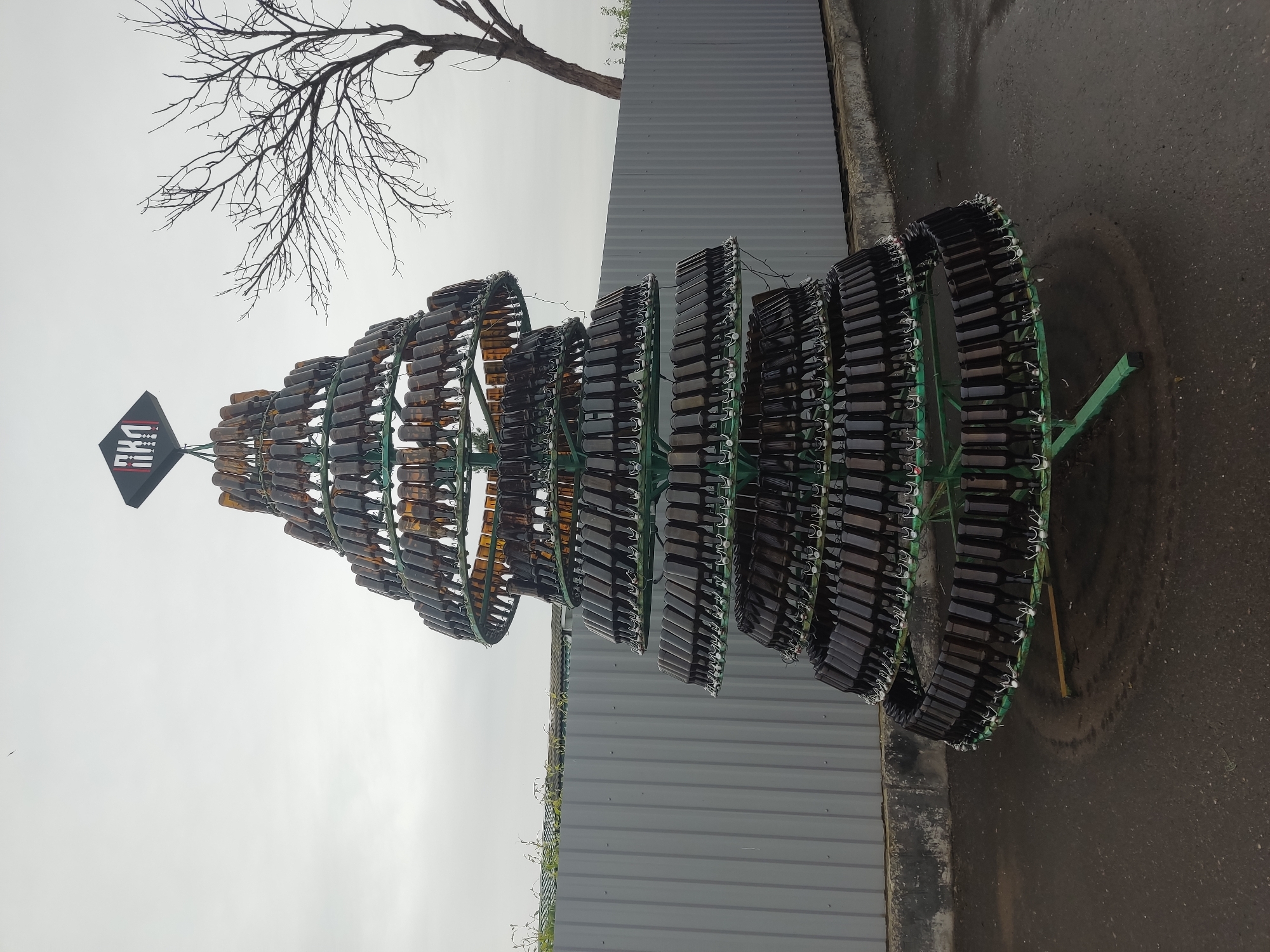 A vertical outdoor shot captures a sculpture in the shape of a Christmas tree made from empty glass bottles in Pskov, Russia. The bottle-tree sculpture features tiered, green metal hoops. Rows of amber and dark-colored bottles encircle each hoop, with the bottle openings facing inward. A black, diamond-shaped sign with red and white designs tops the sculpture. A bare-branched tree stands behind it. The sculpture sits on a gray pavement or asphalt surface next to a gray corrugated metal wall. The sky is overcast.