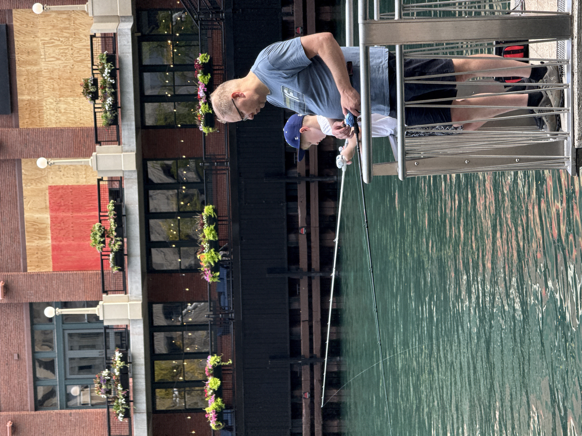 A man and a boy are fishing from a pier in Chicago. The man is wearing a blue t-shirt and glasses. The boy is wearing a blue baseball cap and a white t-shirt. They are both holding fishing rods and looking at the water. The pier is made of concrete and metal railings. In the background, there is a brick building with windows and balconies. There are also some trees and plants. The water is green and reflects the buildings and the sky.