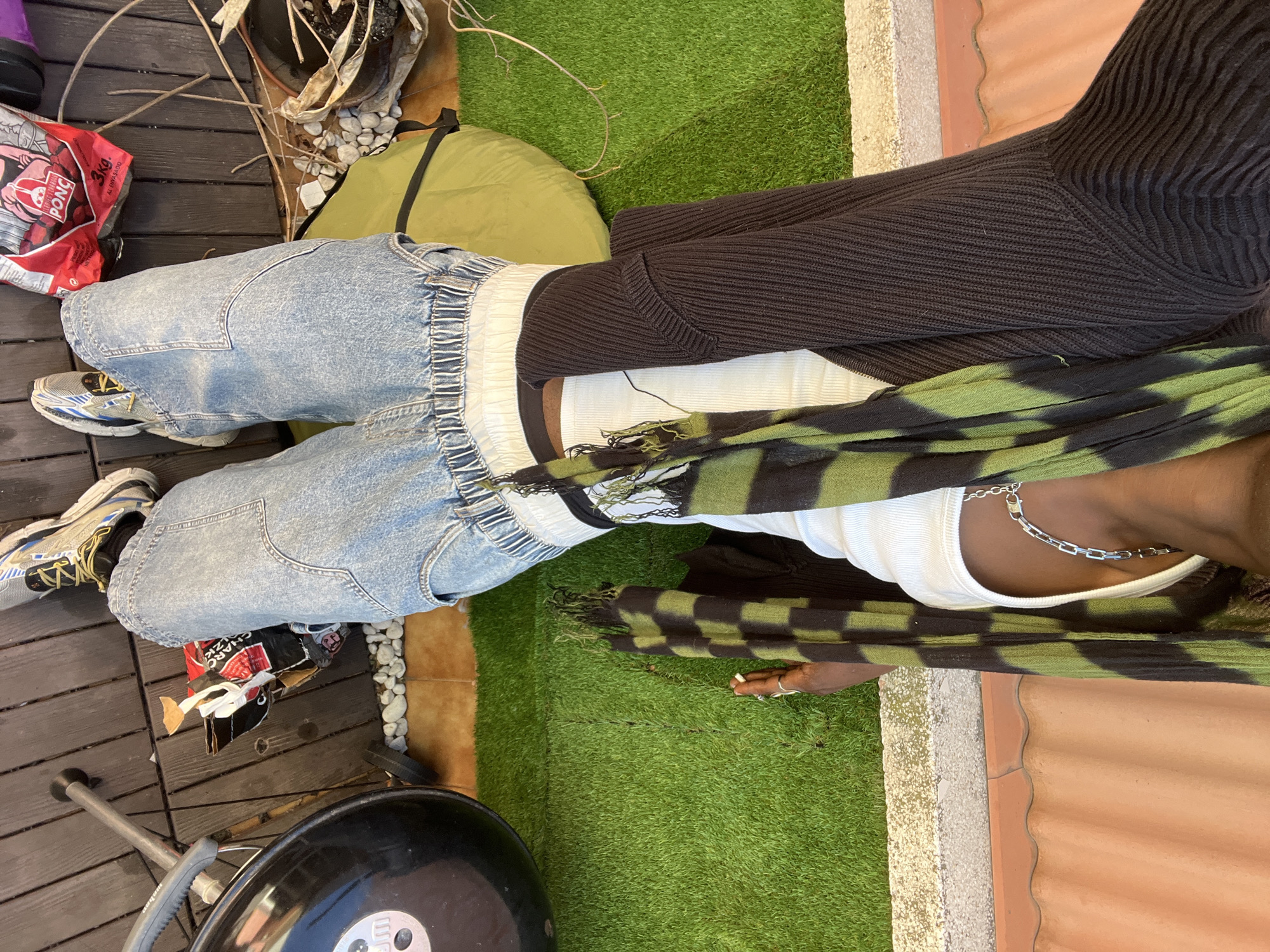 The scene appears to be taken from an overhead perspective on a patio or outdoor area, possibly in a residential setting. The ground covering consists of dark wooden planks, artificial green turf, and terracotta roof tiles.
An individual is positioned in the center of the frame, wearing light blue denim pants with an elastic waistband, a white tank top, and a dark brown sweater draped over the shoulders. A green and black patterned scarf is loosely worn around the neck. A silver chain is visible around the neck.
Various objects are scattered around the area, including a bag of charcoal, a green bag, a grill, stones, sticks and an empty plant pot.