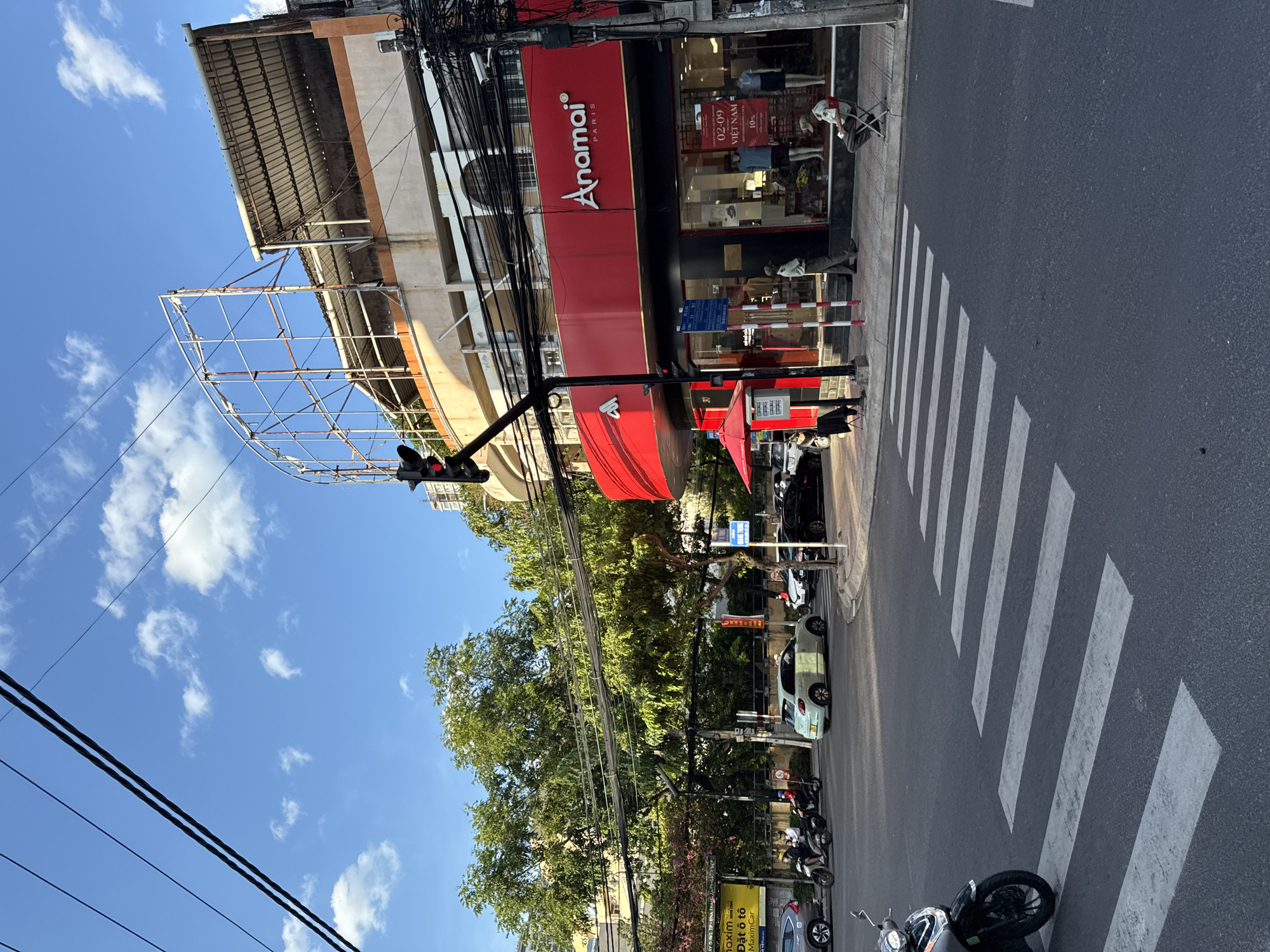 The image depicts a street scene in Nha Trang, Vietnam, characterized by a clear blue sky with scattered white clouds. A prominent red building with the "Anamai PARIS" logo is visible on the right side of the frame, housing a store. In front of the store, mannequins display clothing through large glass windows. A red umbrella is set up on the sidewalk, and a person is seated on a folding chair near the entrance. Across the street, a pedestrian crossing with white stripes is clearly marked on the asphalt. Several vehicles, including motorcycles and cars, are present on the road and at the intersection, suggesting moderate traffic. Tall green trees line the street, partially obscuring some buildings in the background. Overhead, a network of power lines crisscrosses the sky. A traffic light is positioned at the intersection, currently showing a red signal. A derelict billboard structure is visible on top of the Anamai building.