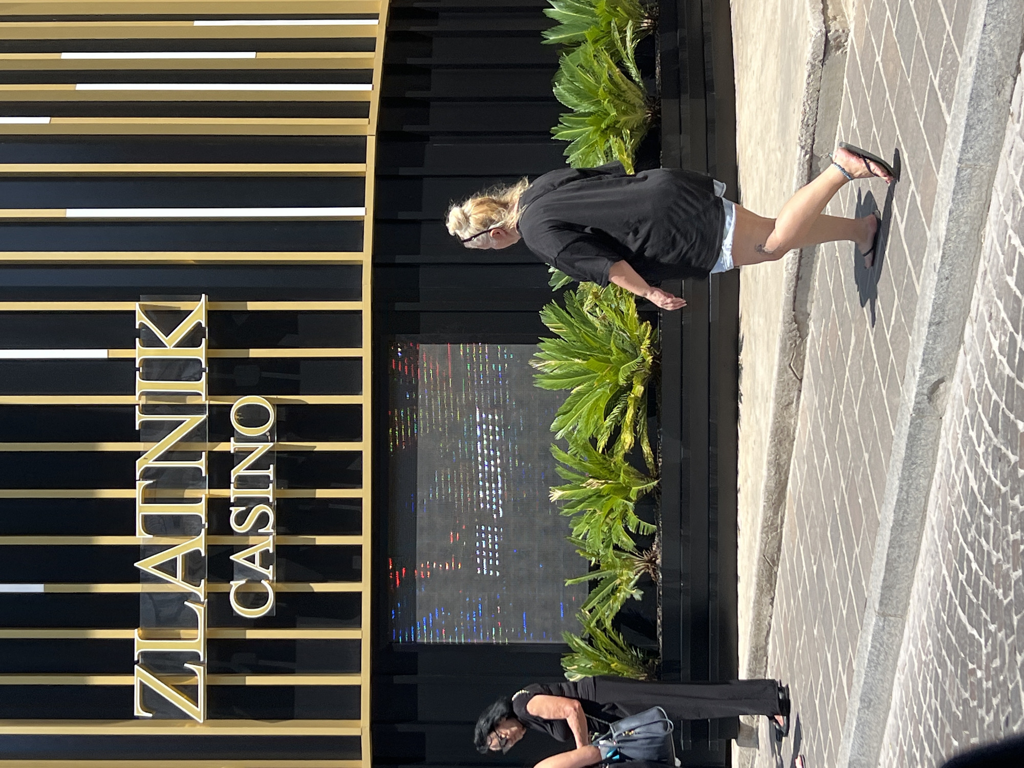 In Budva, Montenegro, in front of the ZLATNIK CASINO, two women walk on a cobblestone street. The woman on the left, with dark hair and sunglasses, wears a black top and pants, carrying a dark blue handbag. The woman on the right, with blonde hair in a bun, is seen from behind, wearing a black t-shirt, denim shorts, and flip-flops. A digital screen displaying a pixelated image is visible behind them, flanked by lush green plants. The casino's facade features vertical golden and black panels.