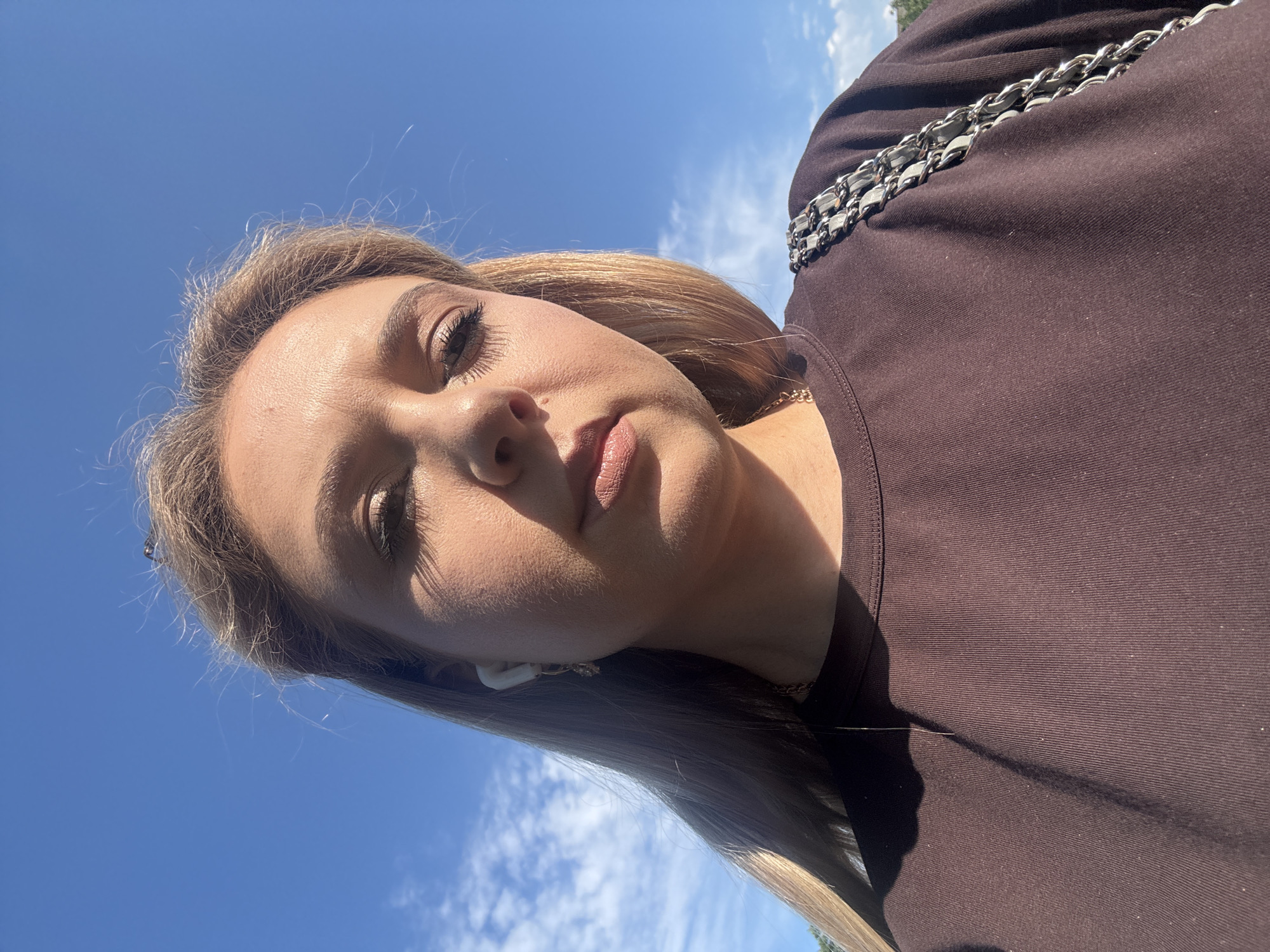 A low-angle, outdoor, medium shot captures a woman from the chest up against a bright blue sky with wispy clouds. She is looking down and slightly to her left, with light blonde hair falling around her shoulders. She is wearing a dark brown crew-neck shirt. A handbag with a silver chain strap is visible on her right shoulder. She is wearing white wireless earbuds in her left ear. A shadow falls across the left side of her face and shirt. The location is Unknown, Unknown.