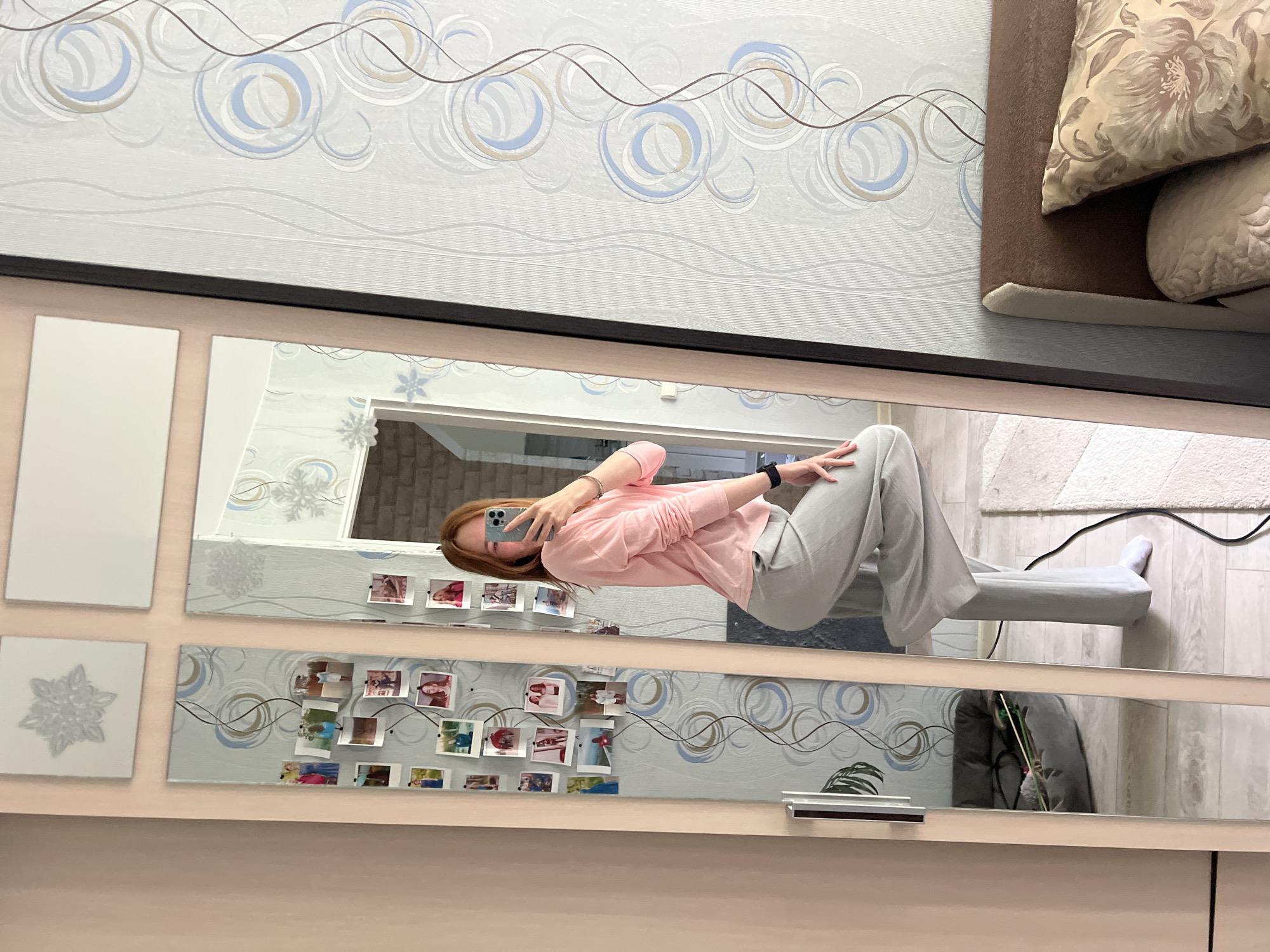 The image is a selfie taken in a room in Chaikovsky, Russia, reflected in a set of decorative mirrors. The woman taking the photo has reddish-brown hair and is wearing a pink long-sleeved shirt and gray sweatpants. She is holding a smartphone in her left hand, presumably to take the photo. The mirrors are mounted on a wall and decorated with patterns and photos. The background includes wallpaper with a circular design, a section of a couch with patterned pillows, a glimpse of the room she is in, including a carpeted floor, and a gray chair or sofa. A black cable runs along the floor.