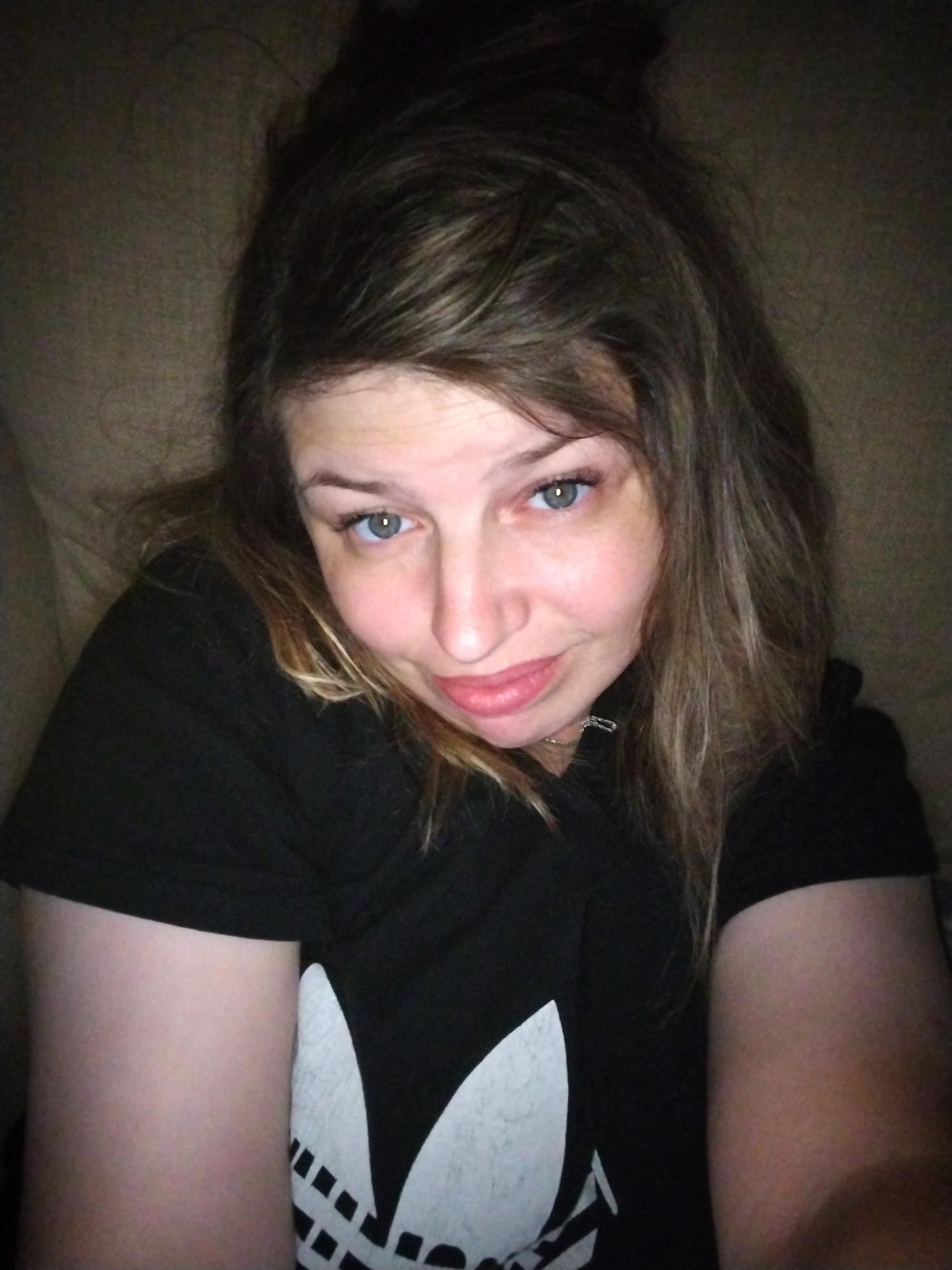 The image is a selfie of a woman with light skin and brown hair. She is wearing a black t-shirt with a white Adidas logo on it. Her hair is pulled up in a messy bun, and she is looking at the camera with a slight smile. The background appears to be a couch or similar upholstered furniture, suggesting an indoor setting. There is no visible information or context within the image to indicate a specific location in the city of Yaroslavl, Russia.