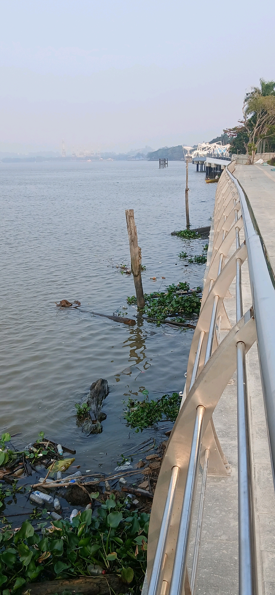 The image shows a view of the Mahakam River in Samarinda, Indonesia, with a walkway in the foreground and a city skyline in the distance. The walkway is made of concrete and has a modern stainless steel railing. The river is calm and appears to be a bit polluted, with some debris visible along the bank and floating on the water, including plastic bottles and water hyacinths. In the distance, there is a faint outline of buildings and a bridge, obscured by haze. There are no people visible in the image. The overall atmosphere is subdued, likely due to the haze and the overcast sky.