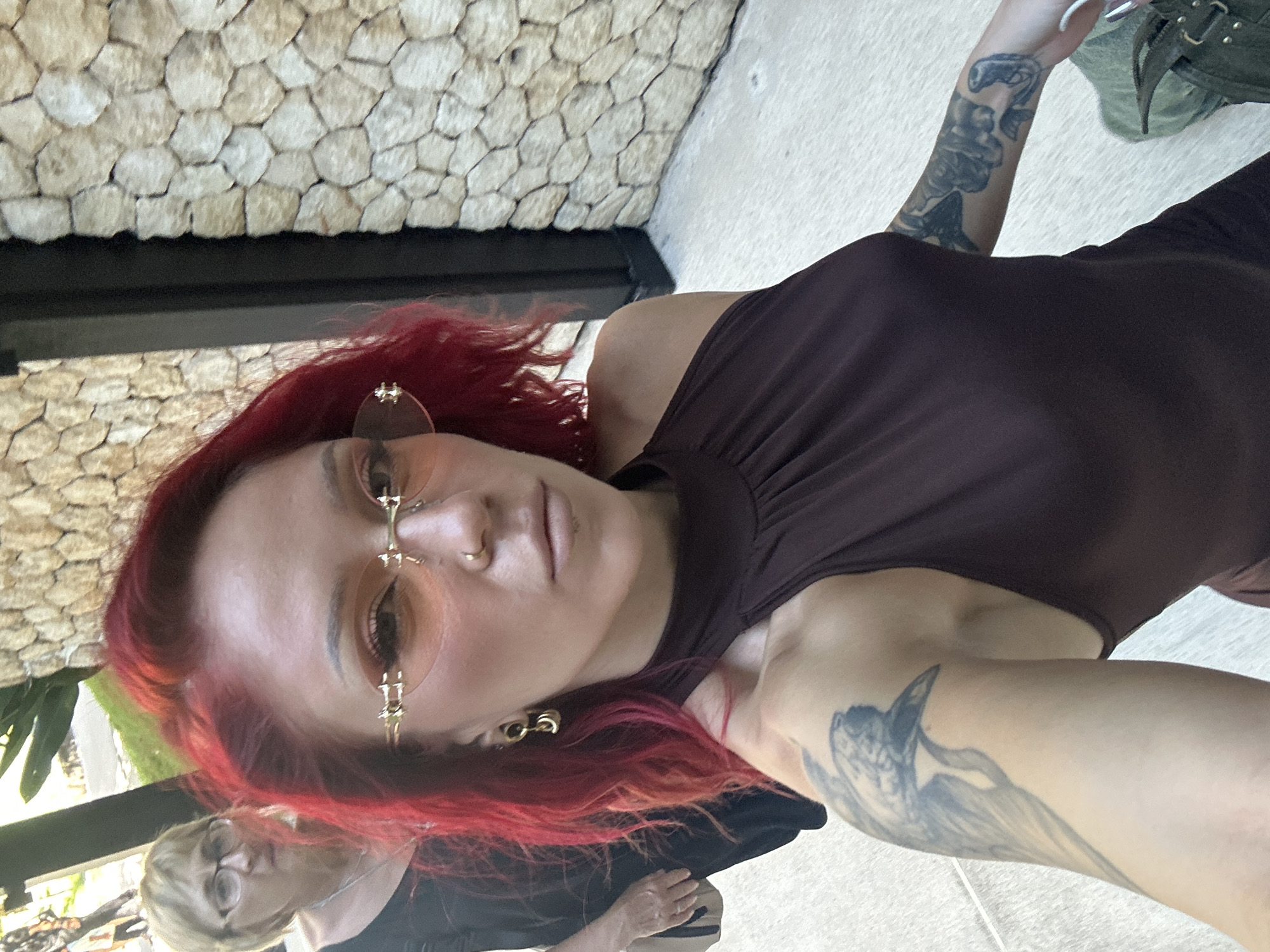 A medium shot of a woman with vibrant red hair and pale skin. She is wearing peach-tinted sunglasses and a dark brown tank top. Her left arm, visible on the right of the frame, has a tattoo. Another tattoo is visible on her right arm on the upper left of the frame. In the lower left corner, another person with blonde hair and glasses is partly visible. The background shows a stone wall and greenery. The location is presumed to be Kuta Selatan, Indonesia.