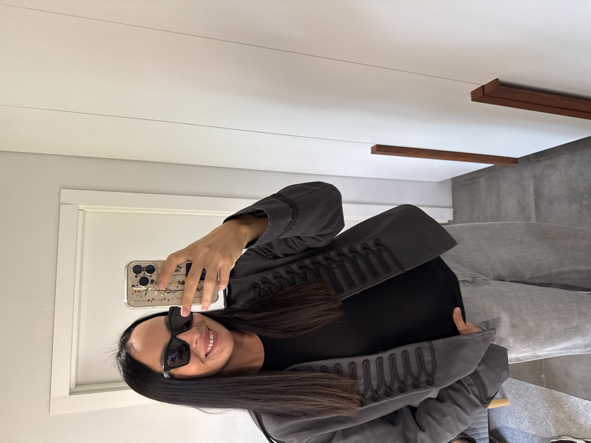 A woman with long, dark hair, wearing black sunglasses and a grey jacket with ornate black detailing, stands in a room and takes a selfie with her phone. She is wearing a black top and light grey jeans. The background shows a white doorframe to her left and a white wardrobe with wooden trim to her right. The flooring appears to be concrete. There is no indication of the city of Chaikovsky, Russia in the image.