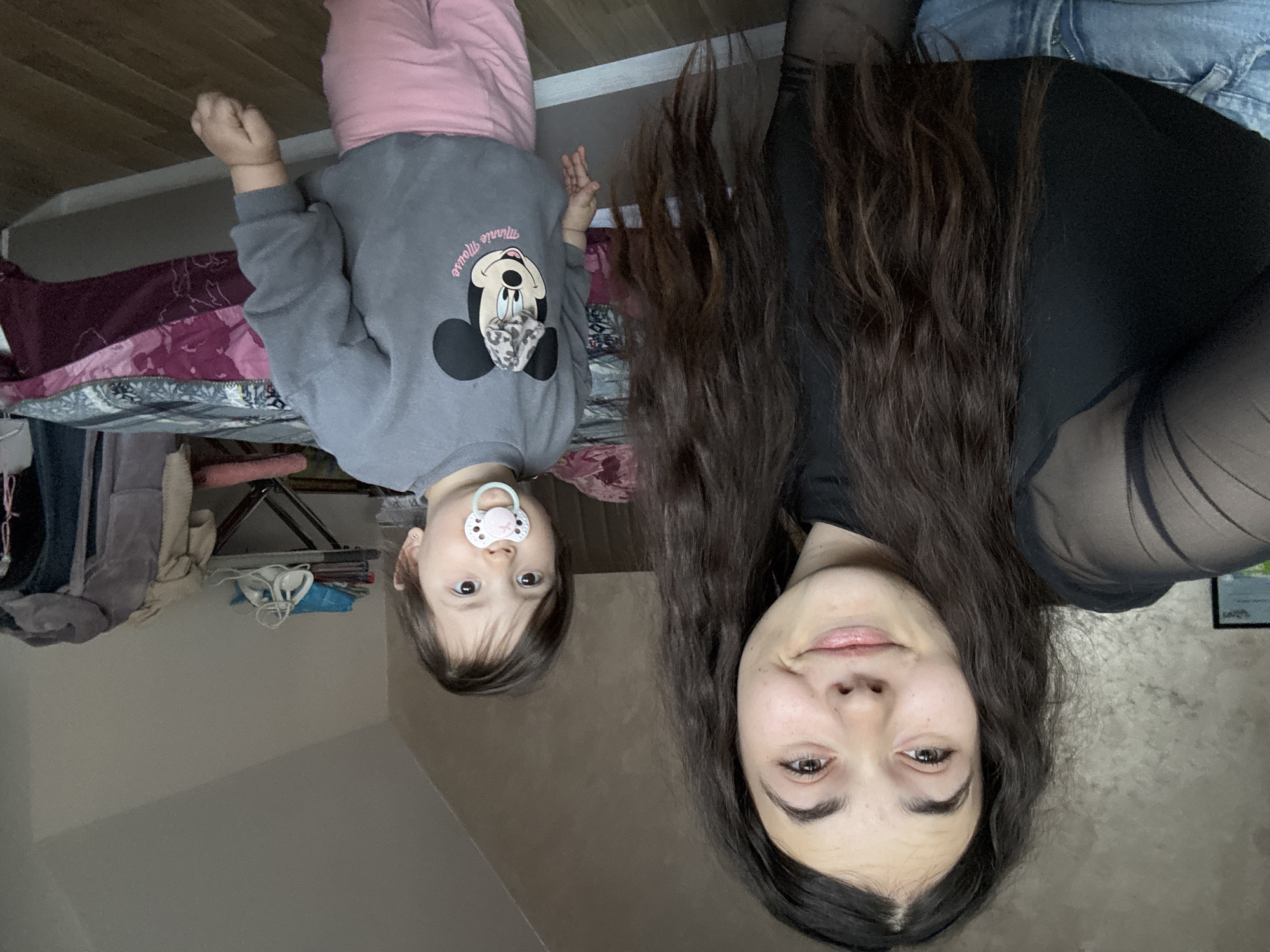 A woman with long, dark brown hair and wearing a black top is positioned to the right of a young child with a pacifier in their mouth. The child is wearing a grey sweatshirt with a Minnie Mouse graphic and pink pants. They appear to be together in an indoor setting, likely a home, as evidenced by a bed with bedding, clothing hanging in the background, and a textured wall. There is no indication of a location in the city of Plovdiv, Bulgaria.
