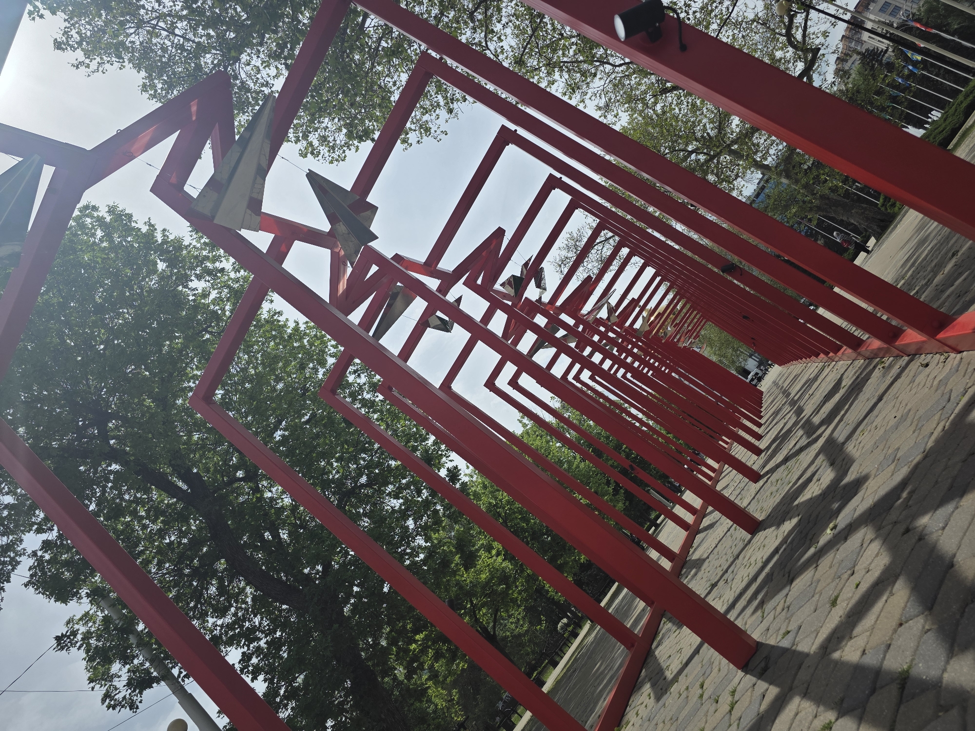 A low-angle shot depicts a long row of red, rectangular frames extending into the distance along a paved pathway. Suspended within the frames are metallic, angular shapes resembling stylized birds or arrows. Trees with green foliage flank the pathway on either side. In the background, flags are visible in front of a building. The location is likely Krasnodar, Russia.