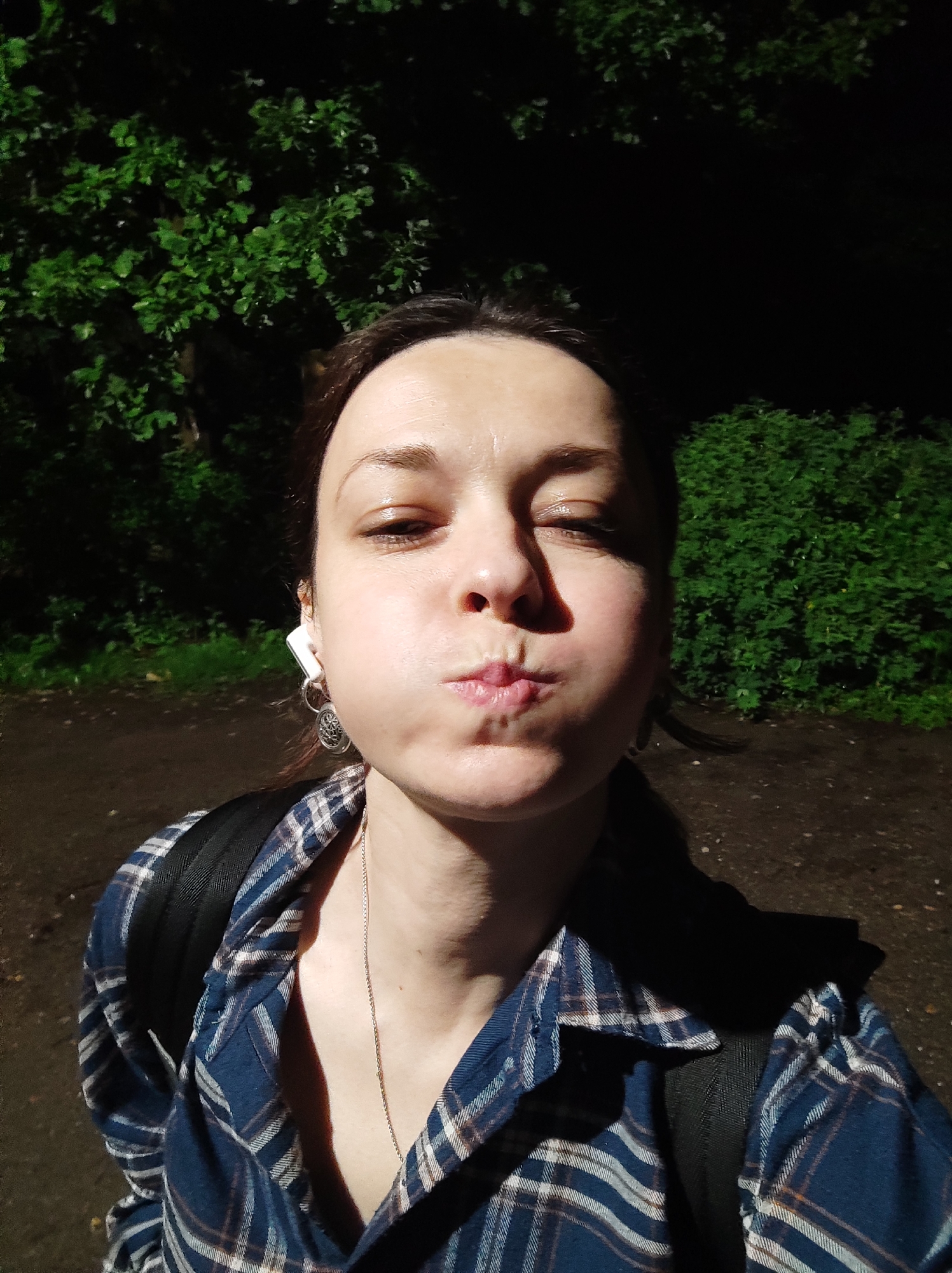 The image is a first-person selfie taken outdoors at night in a wooded or park-like area, possibly in Alabino, Russia. The subject, a woman with her cheeks puffed out and lips pursed, is wearing a blue and white plaid shirt and a backpack. She has white earbuds in her ears, one of which is visible with an earring attached. The lighting suggests an artificial light source is directed at her face, creating a shadow on the left side. The background consists of dark green foliage and a dirt path.