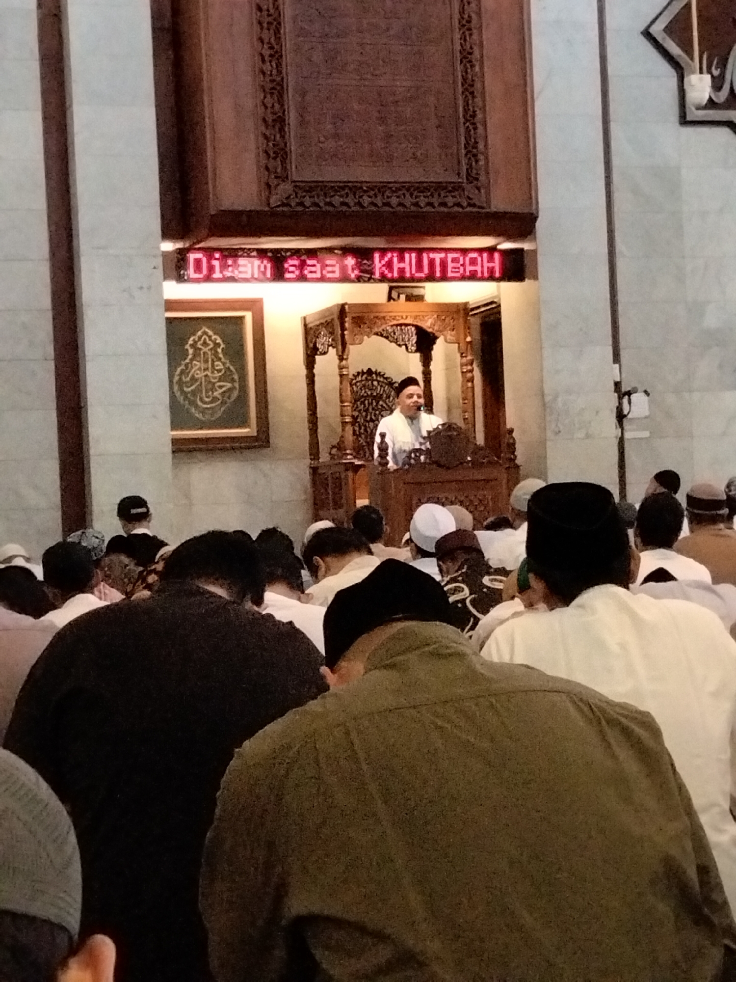 The image depicts a religious gathering inside a mosque, likely in Depok, Indonesia. A man, presumably an imam or preacher, stands at a wooden podium delivering a sermon. He is dressed in a white robe and a white skullcap. Above him, a red LED display reads "DIAM SAAT KHUTBAH" which translates to "SILENCE DURING SERMON." A congregation of men is seated or kneeling before him, facing the podium. They are mostly wearing traditional Islamic attire, including skullcaps and robes. The mosque interior features ornate wooden carvings and a framed religious artwork on the wall. The lighting suggests it is either evening or an indoor setting with artificial illumination. The perspective is from behind the congregation, looking towards the front of the mosque.