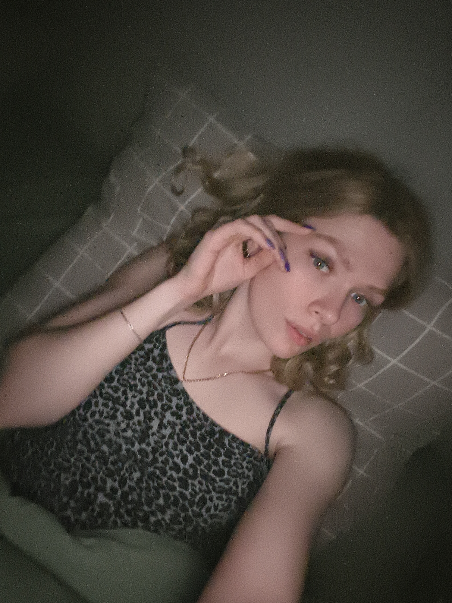 A young woman with light skin and blonde, curly hair is lying on a checkered surface, possibly a bed. She is wearing a leopard print camisole and a gold necklace. She has a bracelet on her left wrist. Her right hand is raised near her face, with her fingers slightly spread. She has painted fingernails. The lighting is dim. There is no information to suggest this image was taken in Русский Вожой, Russia. The location is unknown.