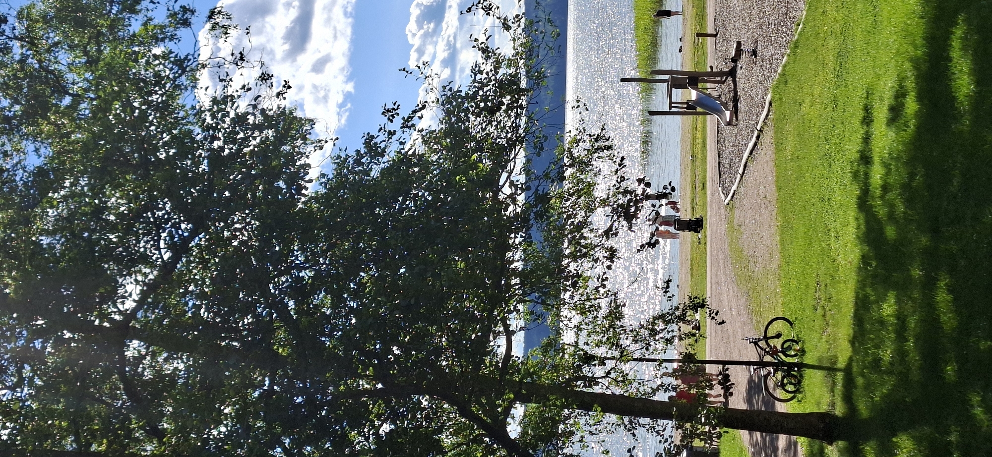 This media file captures a scene by a glistening body of water, likely a lake, on a sunny day in Lahti, Finland. The foreground is dominated by the lush green canopy of a large tree, its branches casting dappled shadows on a grassy area. To the right of the tree, a gravel path winds its way along the water's edge.
Several people are visible in the scene, suggesting a recreational setting. Near the water, a group of individuals are enjoying the day, with some appearing to be in shallow water. Further along the path, a playground structure with a slide is visible, indicating a park or public area. A single bicycle is parked by a tree, leaning against its trunk, suggesting a mode of transportation or a recreational activity.
The water is brightly illuminated by the sun, creating a sparkling effect. In the background, a forested hillside rises from the far shore of the lake. The sky above is a vibrant blue, punctuated by fluffy white clouds, indicative of fair weather. The overall impression is one of a pleasant, leisurely day spent outdoors in a natural setting, with elements of both relaxation and play. The time of day appears to be afternoon, given the angle of the sunlight and the shadows. There is no visible text in the image.