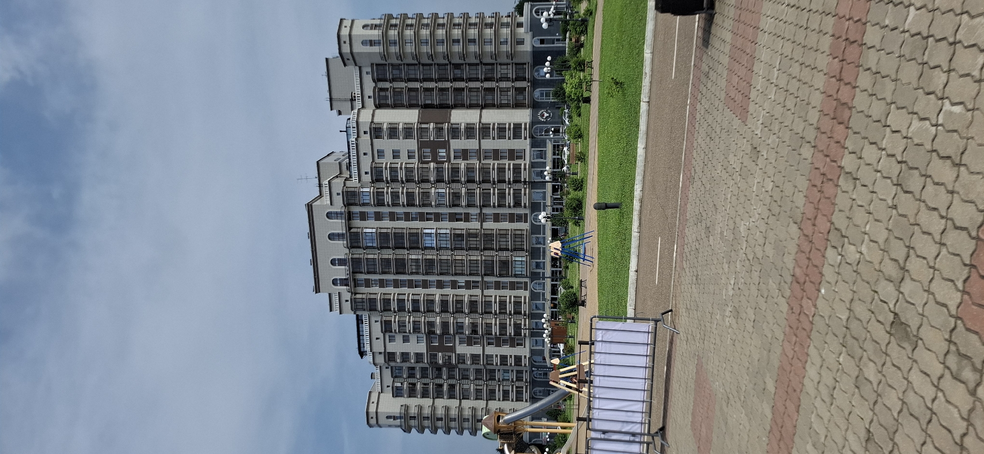 A large, modern residential building with multiple stories and balconies dominates the scene, set against a cloudy sky. In front of the building, a playground area features a slide and swing set. A paved walkway and a grassy area with benches and lampposts are visible in the foreground. The location is the city of Blagoveshchensk, Russia.