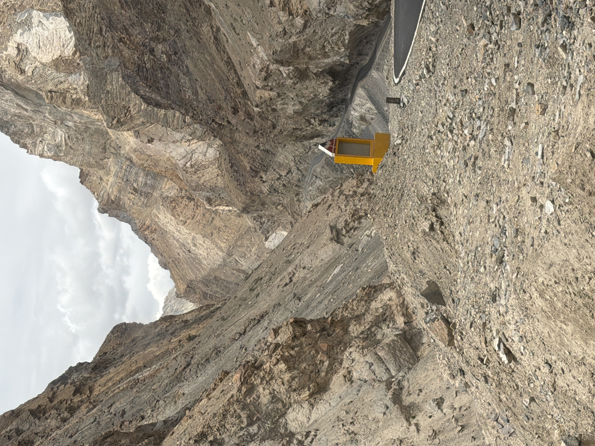 A paved road winds through a narrow, rocky mountain pass. The sides of the pass are steep, with exposed rock formations showing layers of sediment and erosion. A bright yellow structure, possibly a signpost or marker, stands beside the road on the right side, topped with a red and white triangular sign. The ground is covered in loose rocks and gravel. The sky is overcast with scattered clouds. This scene is located near Stongde, India, a region characterized by its dramatic mountainous terrain and the presence of such roads.