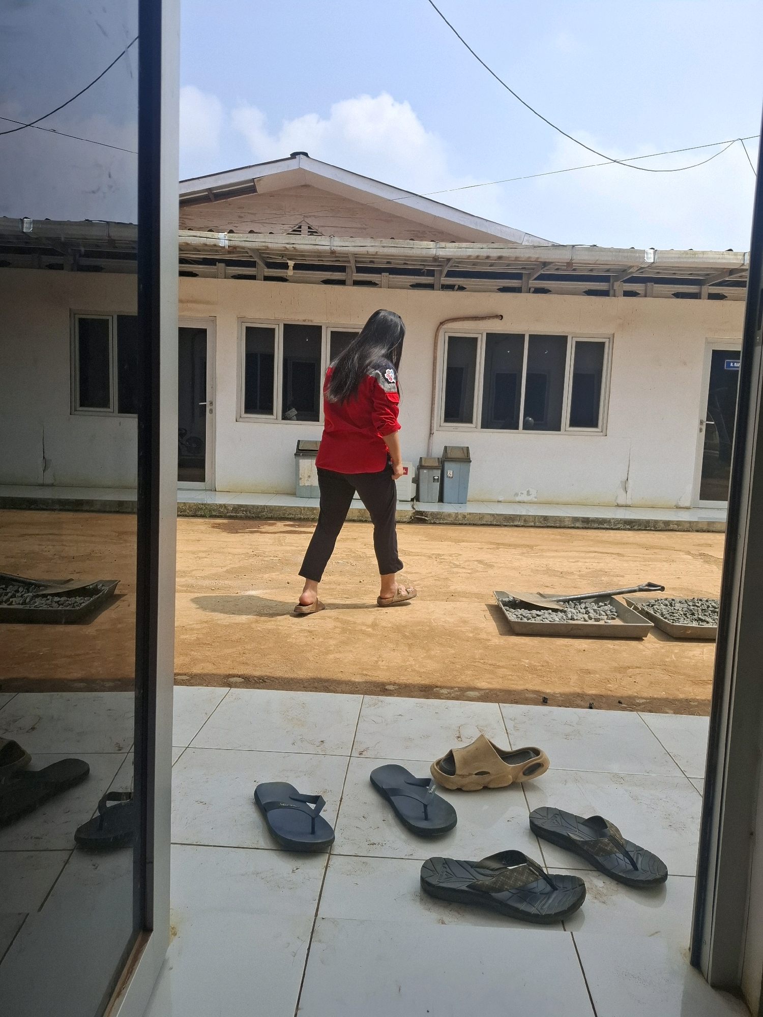 An Indonesian woman with long, dark hair, wearing a red long-sleeved shirt and black pants, walks away from the camera across a sandy courtyard. She is wearing light brown sandals. In the background, a white building with multiple windows and a light-colored roof is visible. Two blue trash cans stand near the building. In the foreground, on a tiled floor, there are several pairs of flip-flops scattered around: one pair of dark blue flip-flops, one pair of dark gray flip-flops, one pair of tan slides, and another pair of dark gray flip-flops. Two shallow trays filled with gravel and a shovel are also present in the courtyard. The scene appears to be in an outdoor area, possibly near a construction site or work area, given the gravel and shovel. The reflection of the building and the sky is visible in the large window or glass door on the left side of the image. The sky is blue with scattered white clouds. The location is identified as Kutasirna, Indonesia.