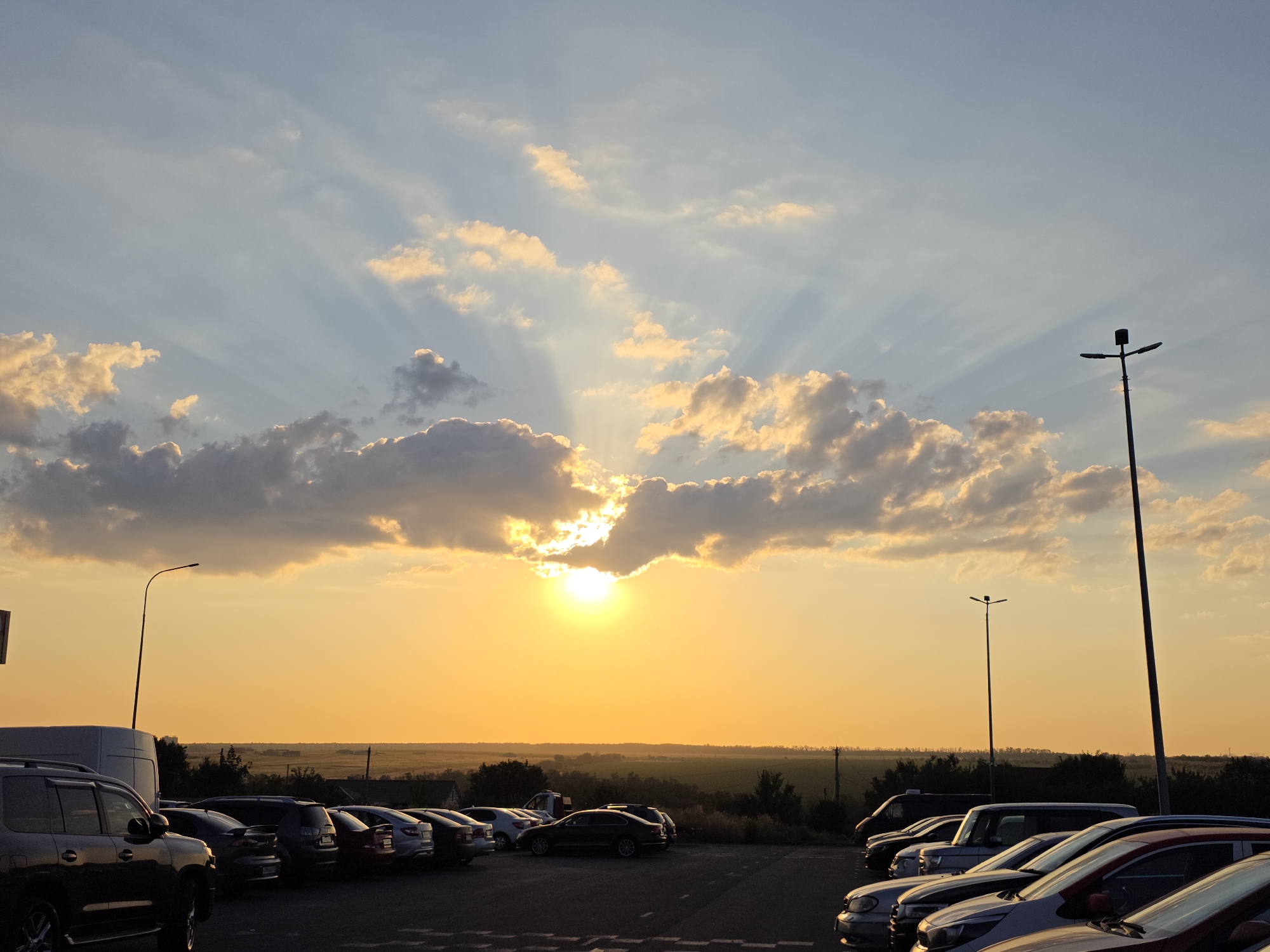 A wide-angle photograph captures a parking lot filled with cars at dusk, with the sun setting in the background. The sky is a gradient of warm oranges and blues, with scattered clouds illuminated by the setting sun, creating a dramatic sunburst effect. The sun itself is partially obscured by a cloud, casting a golden glow over the scene. In the foreground, several cars are parked, their shapes silhouetted against the bright sky. A tall lamppost stands to the right of the frame. The horizon shows a flat landscape with some trees and buildings in the distance. The overall mood is peaceful and serene, indicative of the end of a day. The location is the parking lot of the "Platov International Airport" in Rostov-on-Don, Russia.