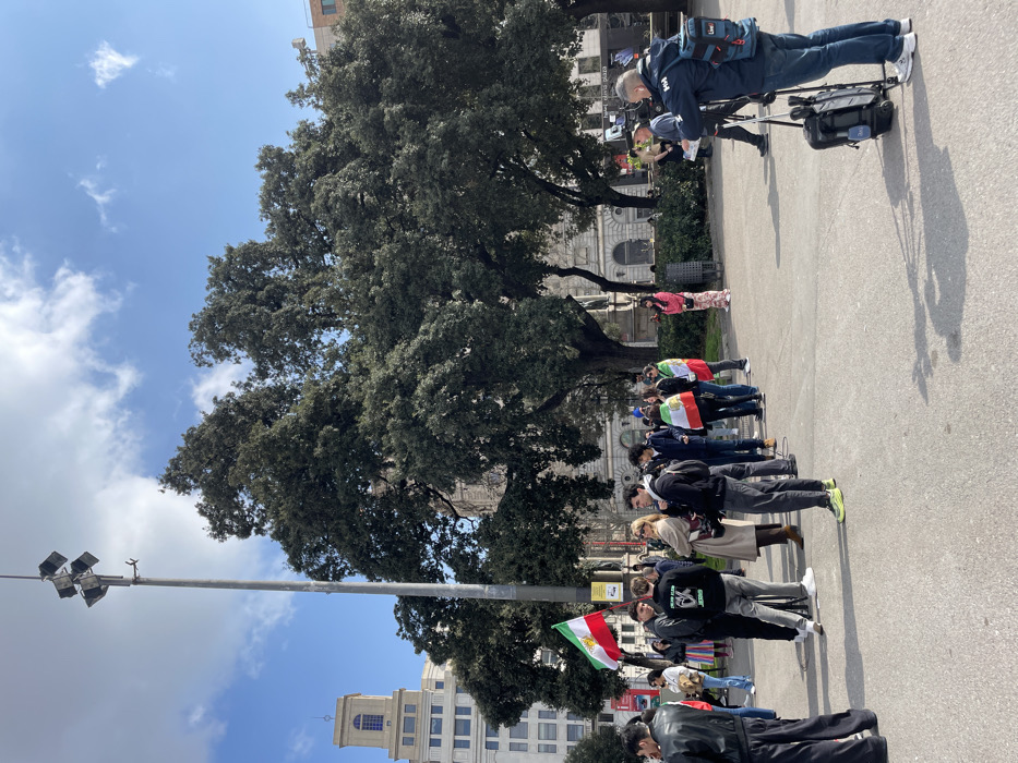 The image depicts an outdoor gathering in Plaça de Catalunya, Barcelona, Spain, under a partly cloudy sky. Approximately two dozen individuals are visible, primarily young adults, assembled on a paved urban square. Several participants are holding or wearing the historical Iranian flag, which features horizontal green, white, and red stripes with a central gold lion and sun emblem.
In the right foreground, a male camera operator wearing a blue jacket with a "Helly Hansen" logo and a blue backpack marked "Live" is positioned behind a professional video camera mounted on a tripod. An equipment cart on wheels is located next to him. Towards the center, a small group of people is engaged in an interaction suggesting an interview, with one individual holding a microphone. A woman in a beige coat and sunglasses observes this interaction.
The background features several large, mature trees with dense, dark green foliage. Behind the trees, classical architectural style buildings are visible. To the far left, a prominent multi-story building with a distinctive tower and a blue-framed window is present. A tall metal pole equipped with multiple floodlights stands near the center-left. Shadows are cast on the ground, indicating direct sunlight.
