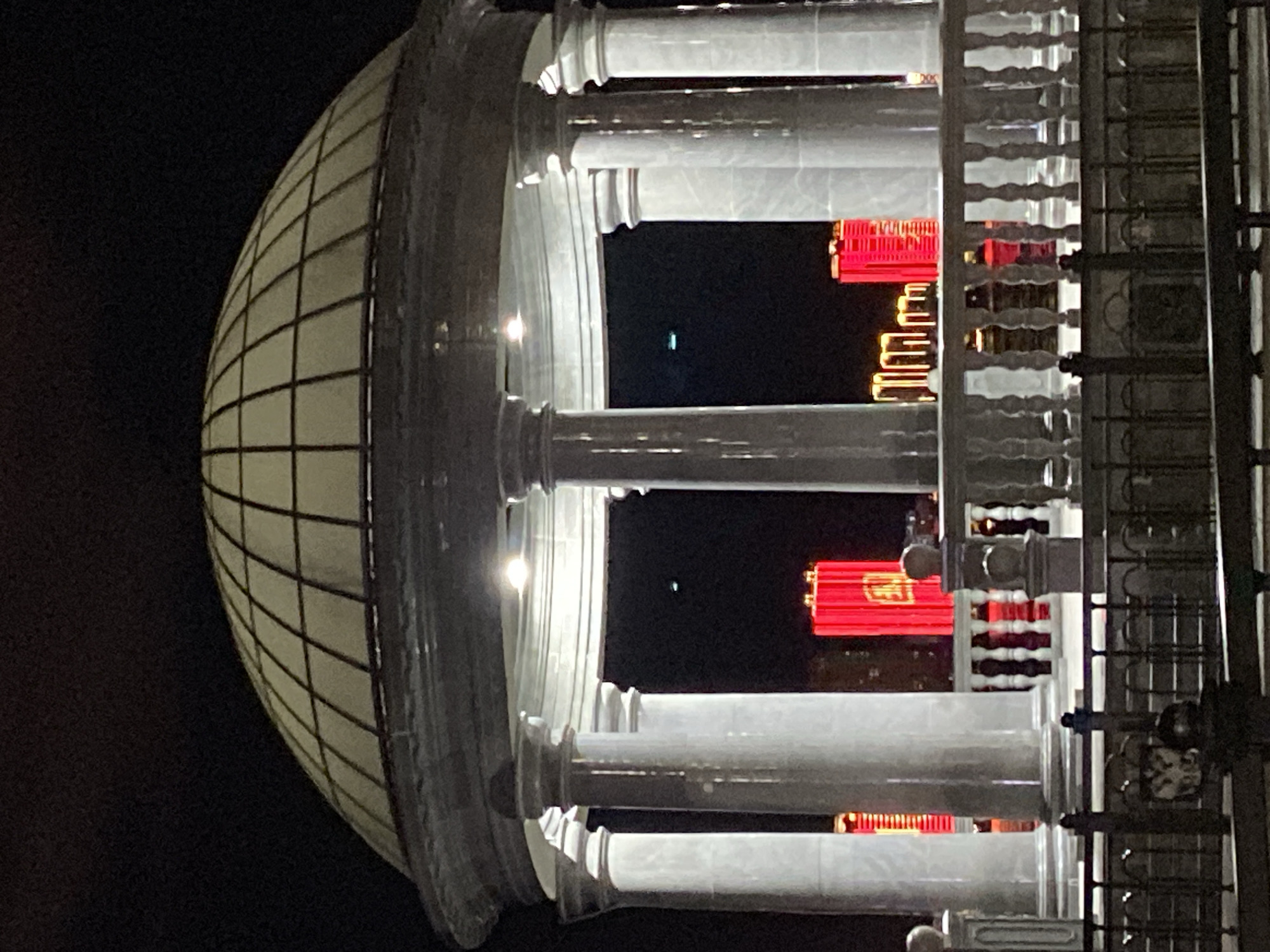 The image captures a nighttime view of a gazebo with a domed roof, supported by classical columns. In the background, illuminated by bright red lights, are modern buildings, likely high-rises, suggesting an urban setting. The gazebo features a white balustrade in front of it, and a dark fence is visible in the foreground. The overall scene appears to be in Blagoveshchensk, Russia, given the presence of lights on buildings in the background that resemble the architecture seen across the Amur River in China, particularly during nighttime illumination displays. There are no people visible in the image.