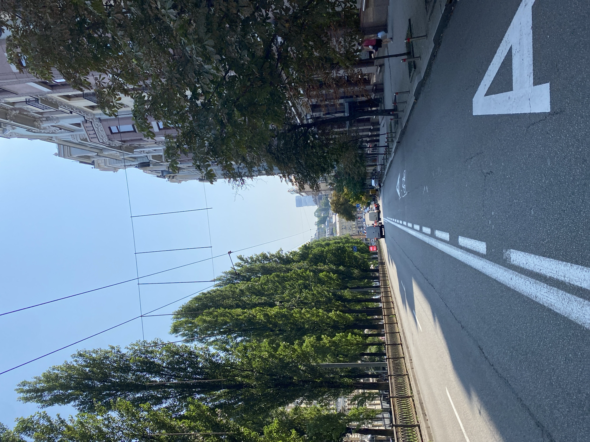 This is a ground-level, wide-angle shot of a city street on a clear, sunny day. The perspective is from the middle of the road, looking down the street. On the left, a row of tall, leafy green trees lines the sidewalk, separated from the road by a decorative metal fence. Across the street to the right, a multi-story building with ornate architecture and balconies rises. The street itself is asphalt, with clear white lane markings. A bicycle lane is indicated by a painted symbol, and a large white number "4" is painted on the road. Several vehicles, including vans and cars, are visible further down the street, suggesting moderate traffic. A few pedestrians are walking on the sidewalk on the right. Overhead, electrical wires crisscross the sky, indicative of a city with trolleybuses or trams. The sky is bright blue with no visible clouds.