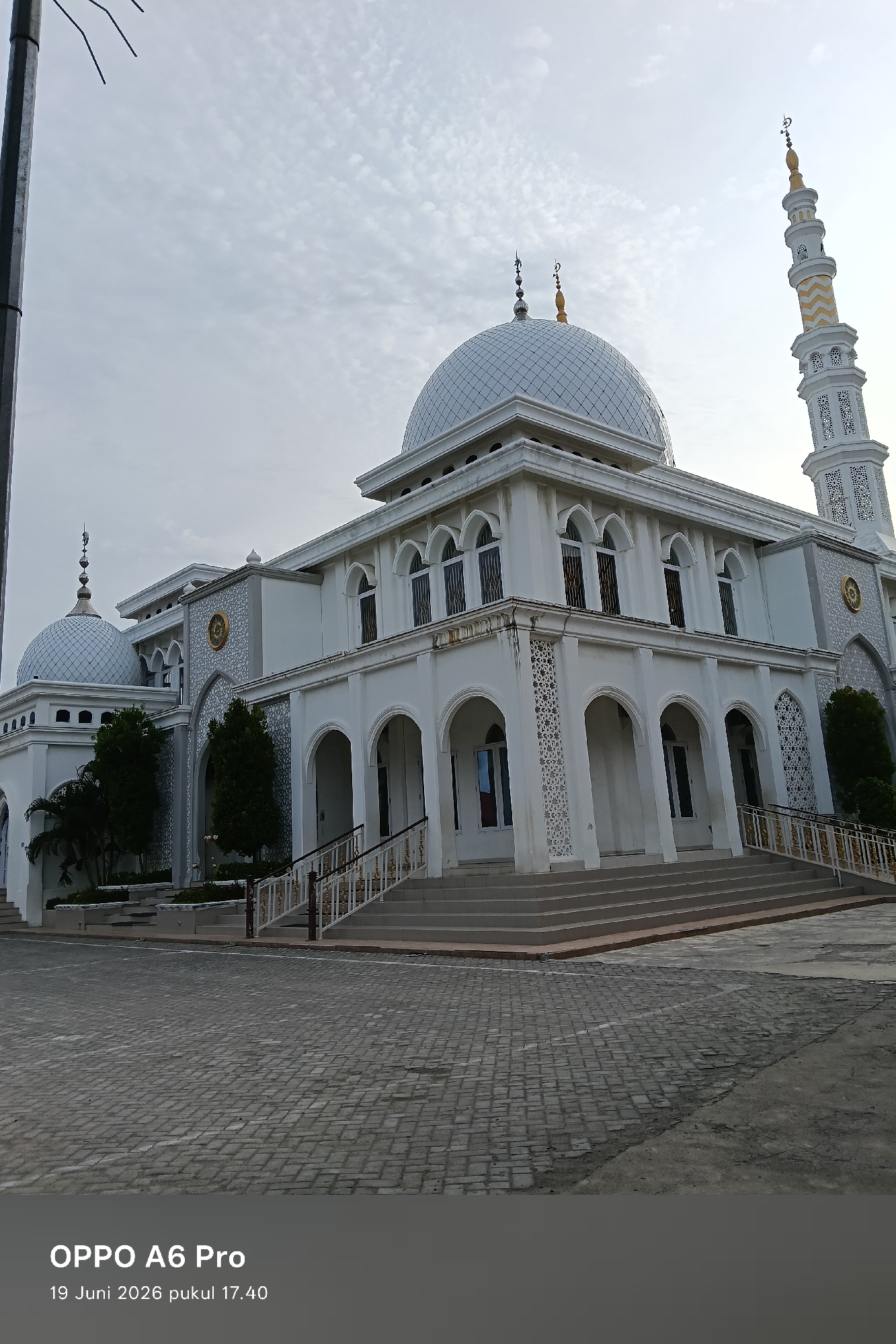 This media file is a photograph of a mosque in Lhokseumawe, Indonesia. The mosque is a grand, white building with multiple domes and a tall minaret. The main structure features a series of arched windows and decorative geometric patterns on its facade. In front of the mosque, there is a wide paved area and a set of stairs leading up to the entrance. Several trees are visible around the building. The sky is overcast, suggesting cloudy weather. The photograph appears to have been taken in the late afternoon, as indicated by the timestamp in the lower corner. The timestamp also shows that the photograph was taken on "19 Juni 2026 pukul 17.40", which translates to June 19, 2026, at 5:40 PM. The image also displays "OPPO A6 Pro", likely indicating the model of the phone used to capture the picture. No people are visible in the photograph.