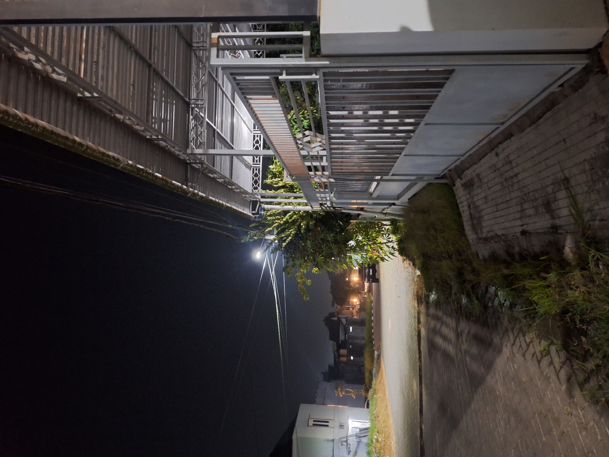 The image depicts an outdoor scene at night in Samarinda, Indonesia. The primary subjects are architectural elements, including a corrugated metal roof, a metal gate, and a brick-paved pathway. Vegetation, such as trees and grass, is also visible, encroaching on the pathway and the metal structures. A street light illuminates the scene, casting long shadows. In the background, houses and a car are discernible, suggesting a residential or semi-residential area. The overall atmosphere is dark and quiet, characteristic of nighttime. The time of day is clearly night, and no specific weather conditions are apparent beyond the darkness. No text is visible in the image.