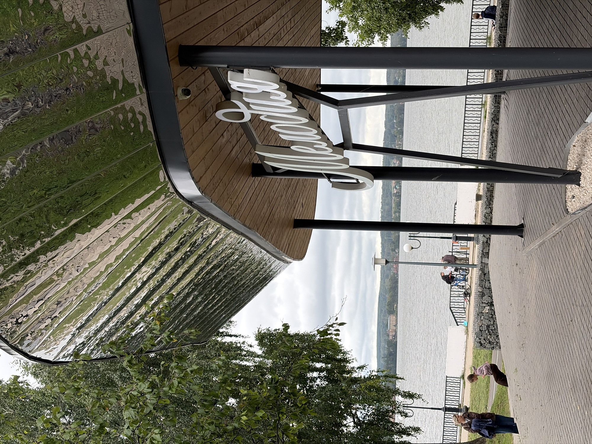 The image shows a modern, curved canopy with a highly reflective, rippled metal surface, supported by dark grey poles. Beneath the canopy, a wooden ceiling is visible, and a large, white, cursive sign reads "Мелодия" (Melodiya). The canopy overhangs a paved area leading down to a body of water, likely a lake or river, with a distant treeline and buildings on the far shore. A black metal fence with an ornate, wavy pattern runs along the waterfront.
In the foreground, a paved walkway and steps are visible. To the left, a woman with a backpack stands on the steps, facing away from the camera and towards the water. A child is in motion on the steps, appearing to be running or jumping down. Further along the waterfront, a group of people are gathered near the fence, one of whom is with a bicycle. In the background, a solitary figure sits on the ground near the water's edge. The sky is overcast and cloudy. The architectural style and signage suggest this is a public space, possibly a cafe, restaurant, or recreational area, located in Votkinsk, Russia, a city known for its association with the composer Pyotr Ilyich Tchaikovsky.