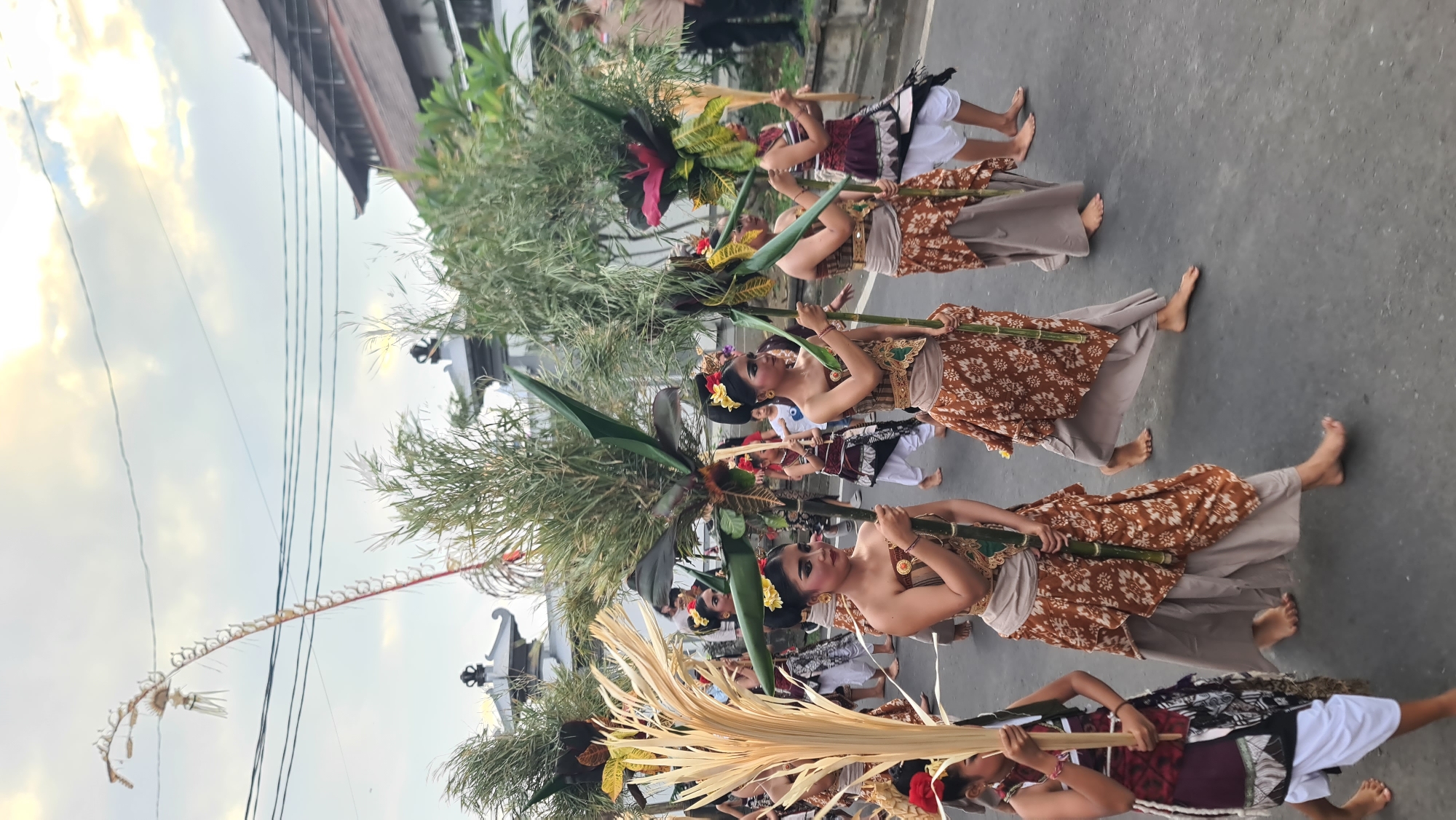 This image captures a traditional Balinese procession or dance performance taking place outdoors, likely in Denpasar, Indonesia. The scene features a group of individuals, primarily women, dressed in elaborate, colorful traditional attire. Their costumes consist of intricately patterned fabrics, often with gold accents and decorative headpieces adorned with flowers. They are holding long, slender poles, some of which are topped with large, leafy green decorations and long, straw-like elements. Some of the dancers are barefoot, a common practice in traditional Indonesian performances.
The procession appears to be moving along a paved road or walkway, with a building or structure with traditional Balinese architecture visible in the background. The sky suggests it is either early morning or late afternoon, with soft, diffused sunlight casting a warm glow on the scene. The weather appears to be pleasant, with a partly cloudy sky.
The overall mood seems festive and ceremonial, indicating a cultural event or celebration. The focused expressions of the dancers suggest they are engaged in a formal performance. The presence of overhead wires and the modern road surface indicate an urban or semi-urban setting, possibly a street parade or a public ceremony. The image is oriented vertically. No visible text is present in the image.