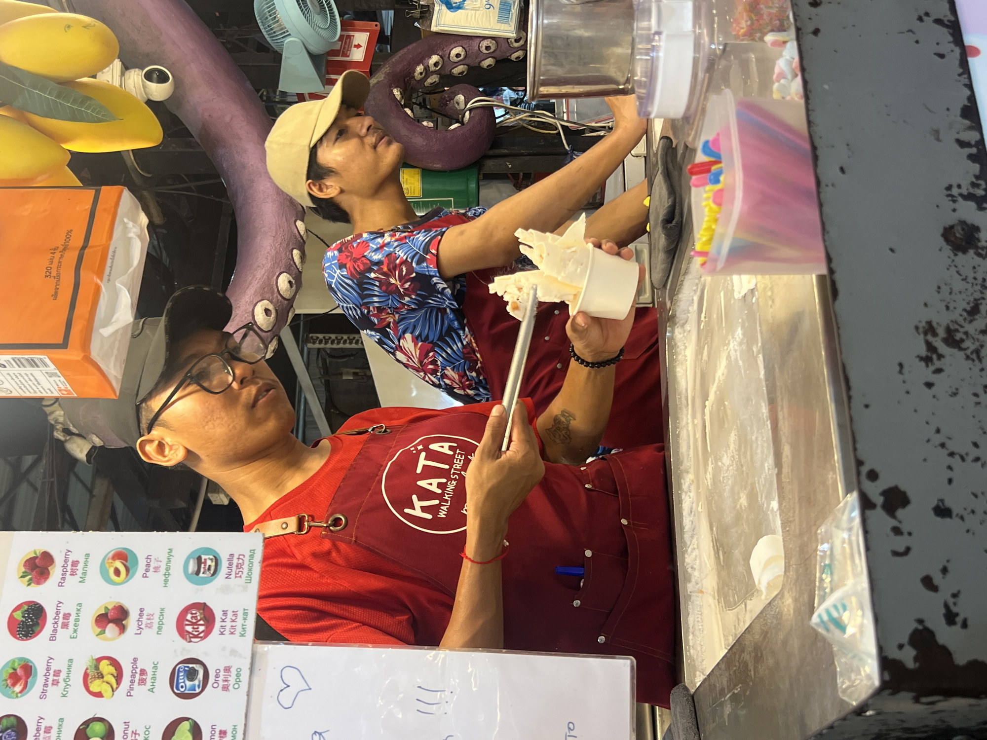 This media file captures a street food vendor in action, likely in Karon, Thailand, as indicated by the menu text. The image features two individuals, appearing to be young men, working at a dessert stall. One man, wearing glasses and a red apron with the "KATA WALKING STREET" logo, is preparing a rolled ice cream. He is holding a metal spatula, scraping and shaping the ice cream on a frozen surface. The other man, wearing a red apron over a colorful floral shirt and a beige baseball cap, is looking upwards, possibly at something off-camera or engaged in conversation.
The stall itself is bustling with activity. Various ingredients and supplies are visible, including a container of colorful sprinkles and what appear to be marshmallows in the background. To the left, a prominent display of fruit illustrations on what looks like a menu board showcases options like strawberries, raspberries, blackberries, lychees, peaches, pineapples, and coconuts, along with branded items like Kit Kat and Oreo, suggesting a wide variety of ice cream flavors and toppings.
In the background, a large, purple, tentacle-like decoration with eye-like details hangs, adding a unique and whimsical element to the scene. A blue fan is also visible, suggesting a warm climate, and the overall impression is of a lively outdoor market or street food setting, likely during daylight hours. The focus on food preparation and the presence of customers (implied by the vendor-staff interaction) point to a busy commercial environment. The overall mood seems energetic and focused on the task of serving customers.